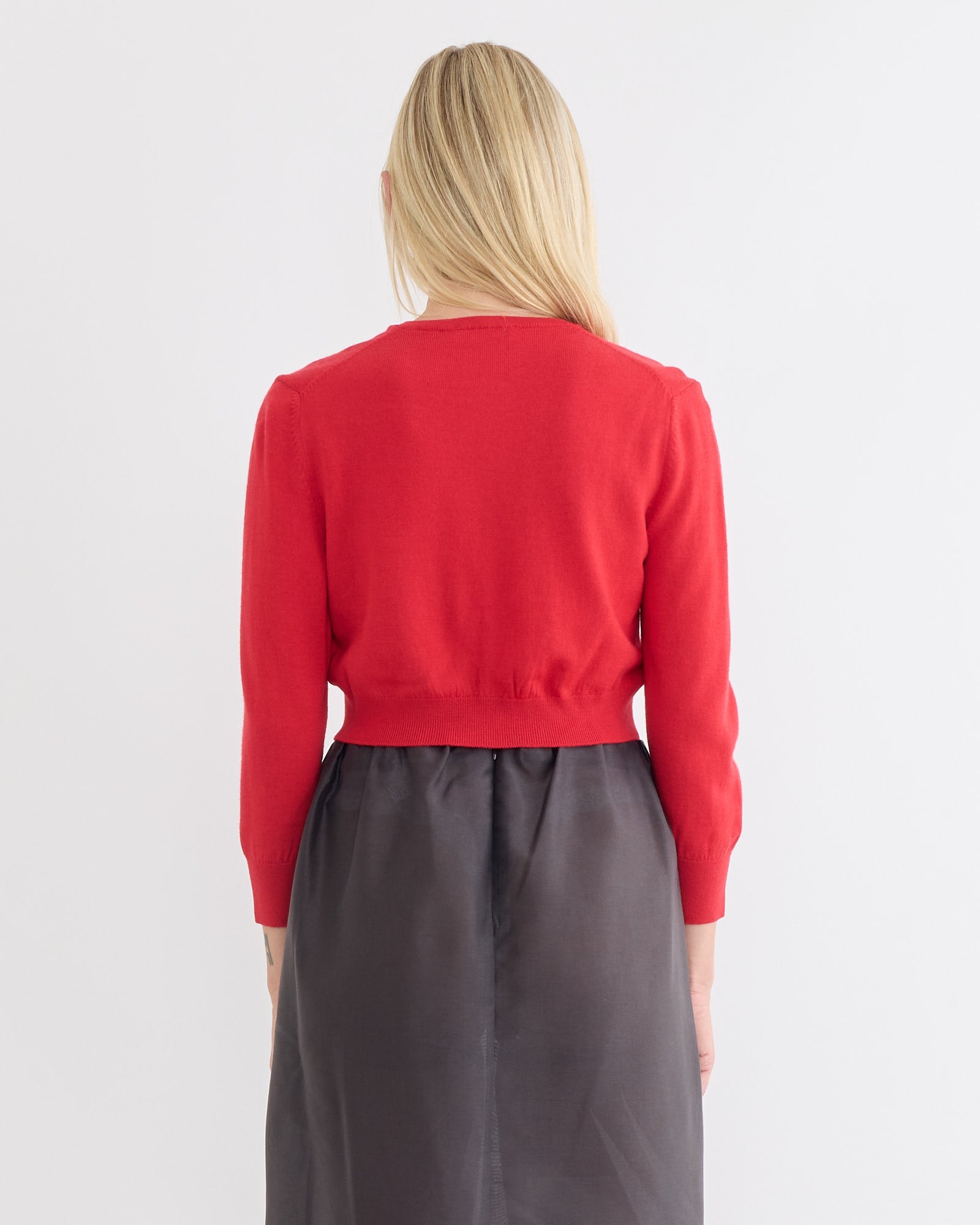 A person with long blonde hair, seen from behind, wears the Amomento Round Neck Crop Cardigan in Red with a high-waisted dark gray skirt, standing against a plain white background.