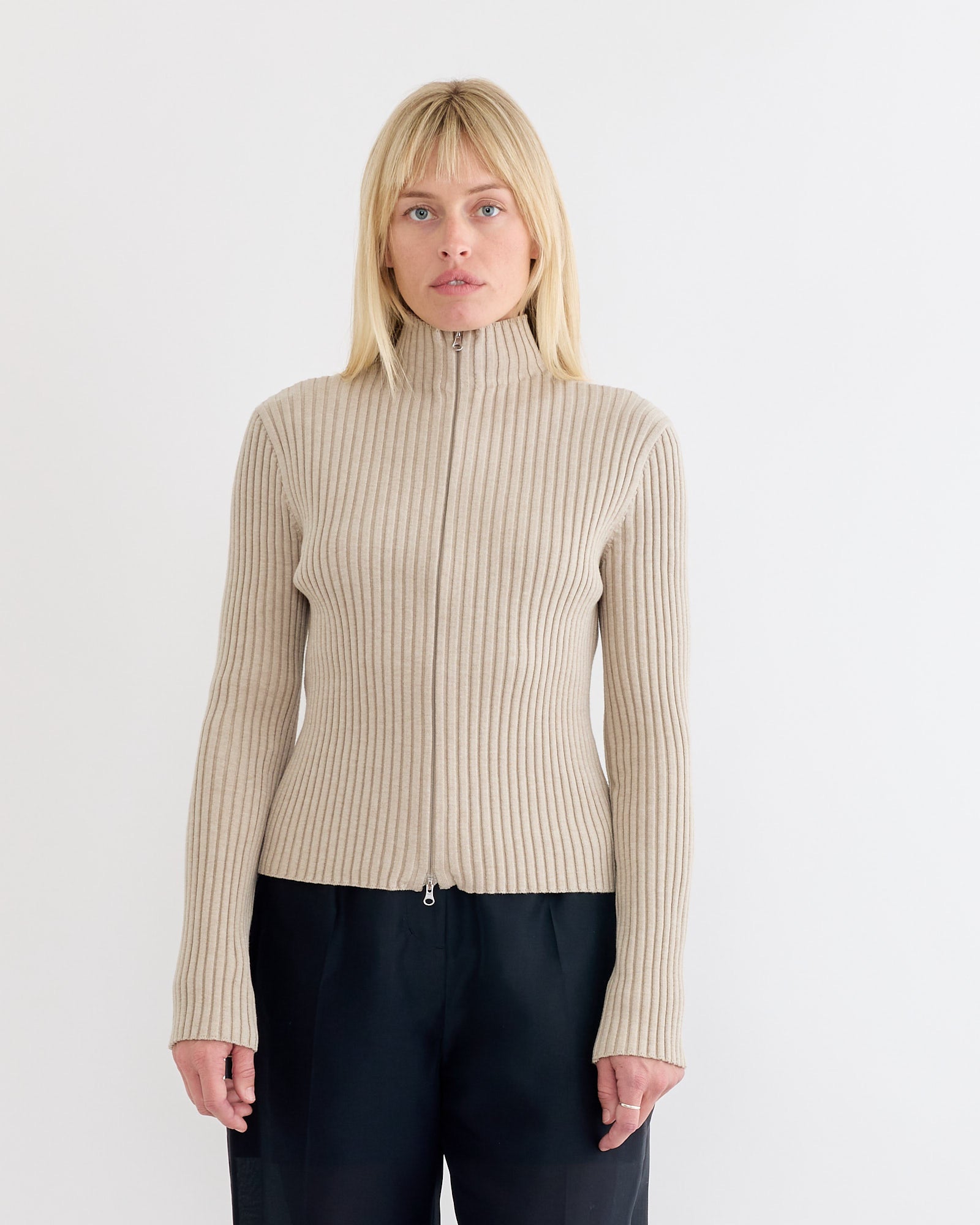 A woman with straight blonde hair and a neutral expression wears the Amomento Ribbed High Neck Zip Up Cardigan in Beige, styled over loose black pants, standing against a plain white background.