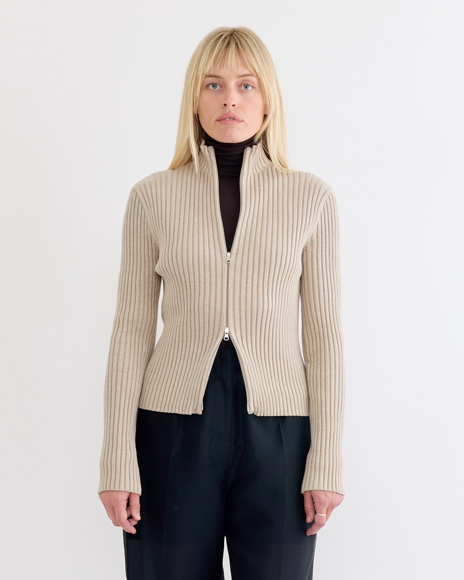 A woman with straight blonde hair wears the Amomento Ribbed High Neck Zip Up Cardigan in Beige over a black turtleneck and black pants, standing against a plain white background.