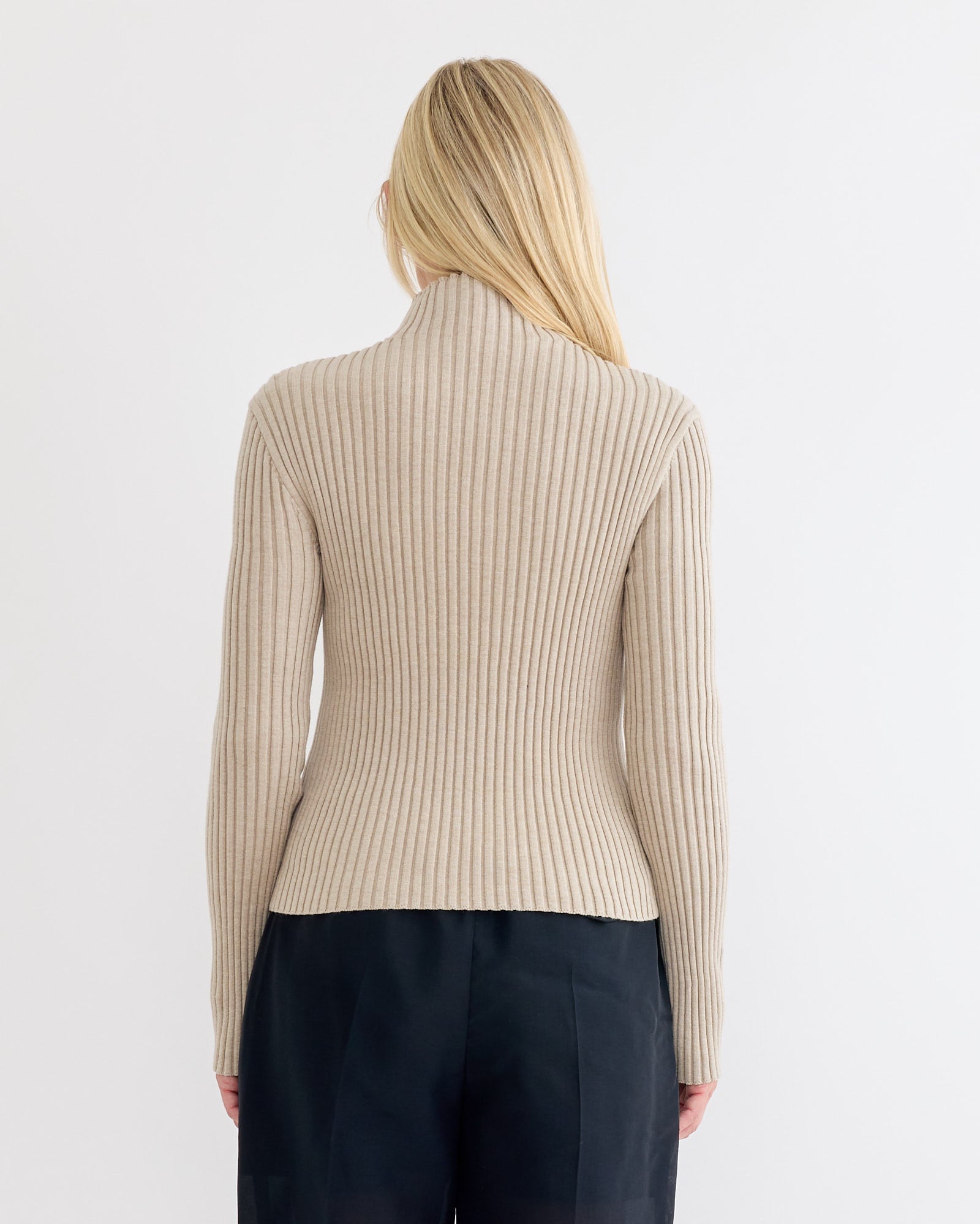 A person with long blonde hair is seen from the back wearing the Amomento Ribbed High Neck Zip Up Cardigan in Beige and black pants against a plain white background.