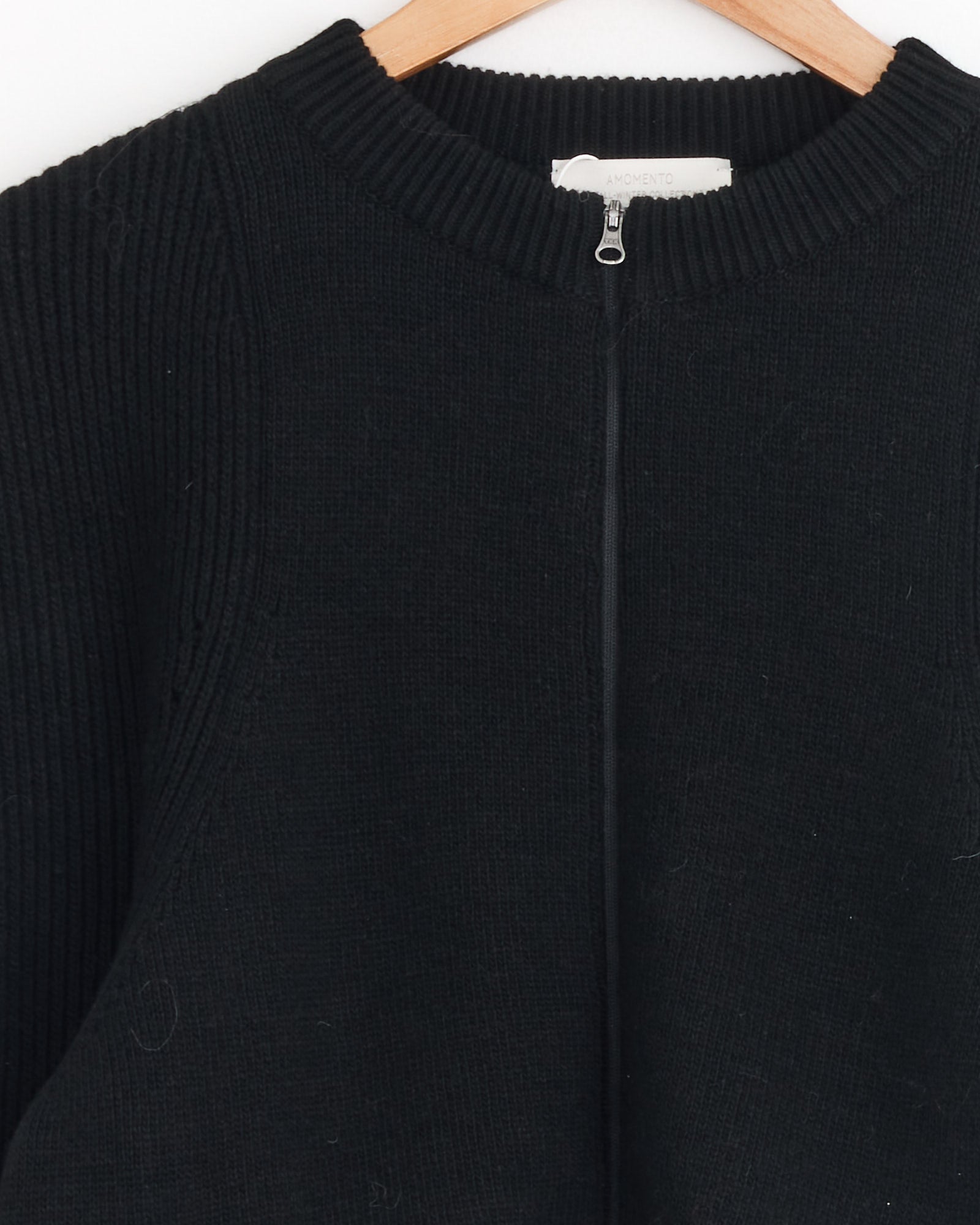 The Cut Out Rounded Cardigan in Black by Amomento features a zip-up front with ribbed collar and shoulders, shown on a wooden hanger against a white background. For sizing and fit questions, contact us through our online chat.
