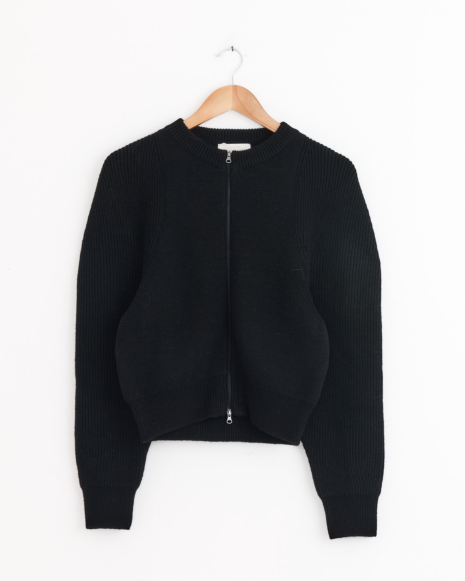 The Cut Out Rounded Cardigan in Black by Amomento, featuring long sleeves and a zip-up ribbed design, hangs on a wooden hanger against a white background. For sizing or fit questions, contact our online chat for assistance.