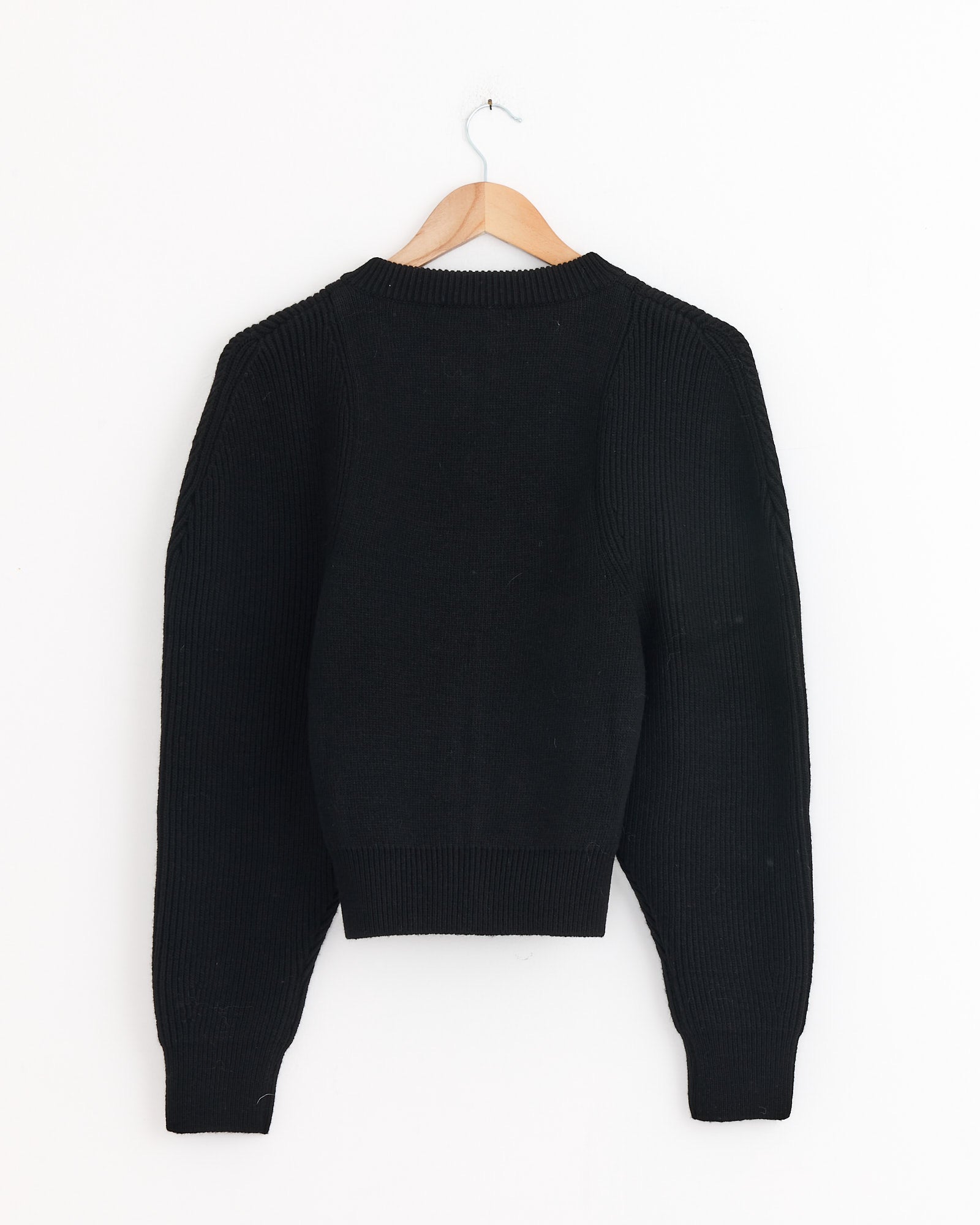 The Amomento Cut Out Rounded Cardigan in Black is displayed with the back facing forward on a wooden hanger against a white wall. For questions about sizing or fit, please use our online chat for assistance.