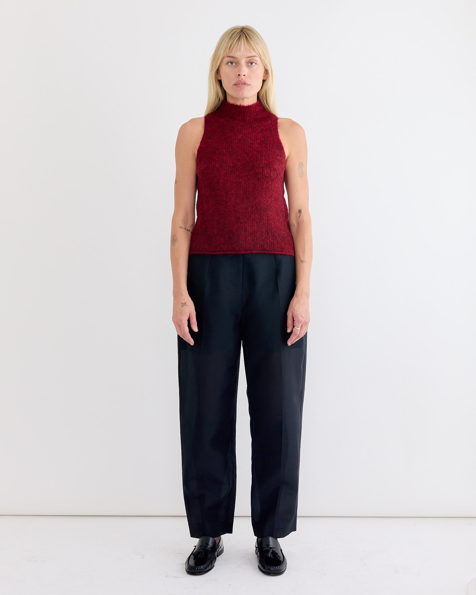 A woman with straight blonde hair stands against a white wall, wearing the Amomento Sheer Sleeveless Top in Red, paired with black wide-leg pants and black shoes. She faces forward, arms relaxed at her sides.