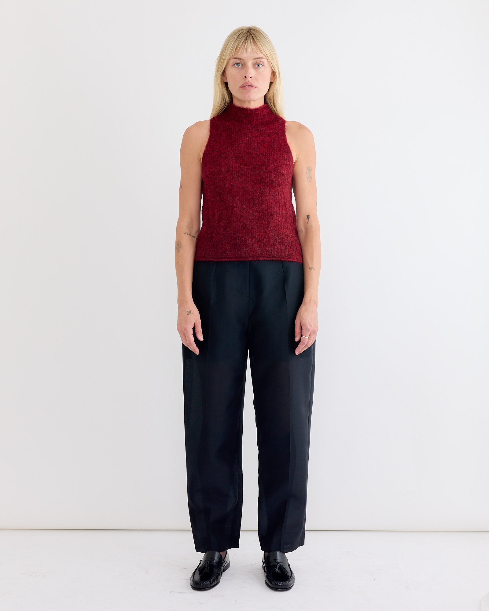 A woman with straight blonde hair and arm tattoos stands against a white wall, wearing Amomento’s Organza Structure Pant in Black, a sleeveless high-neck burgundy top, and black shoes.