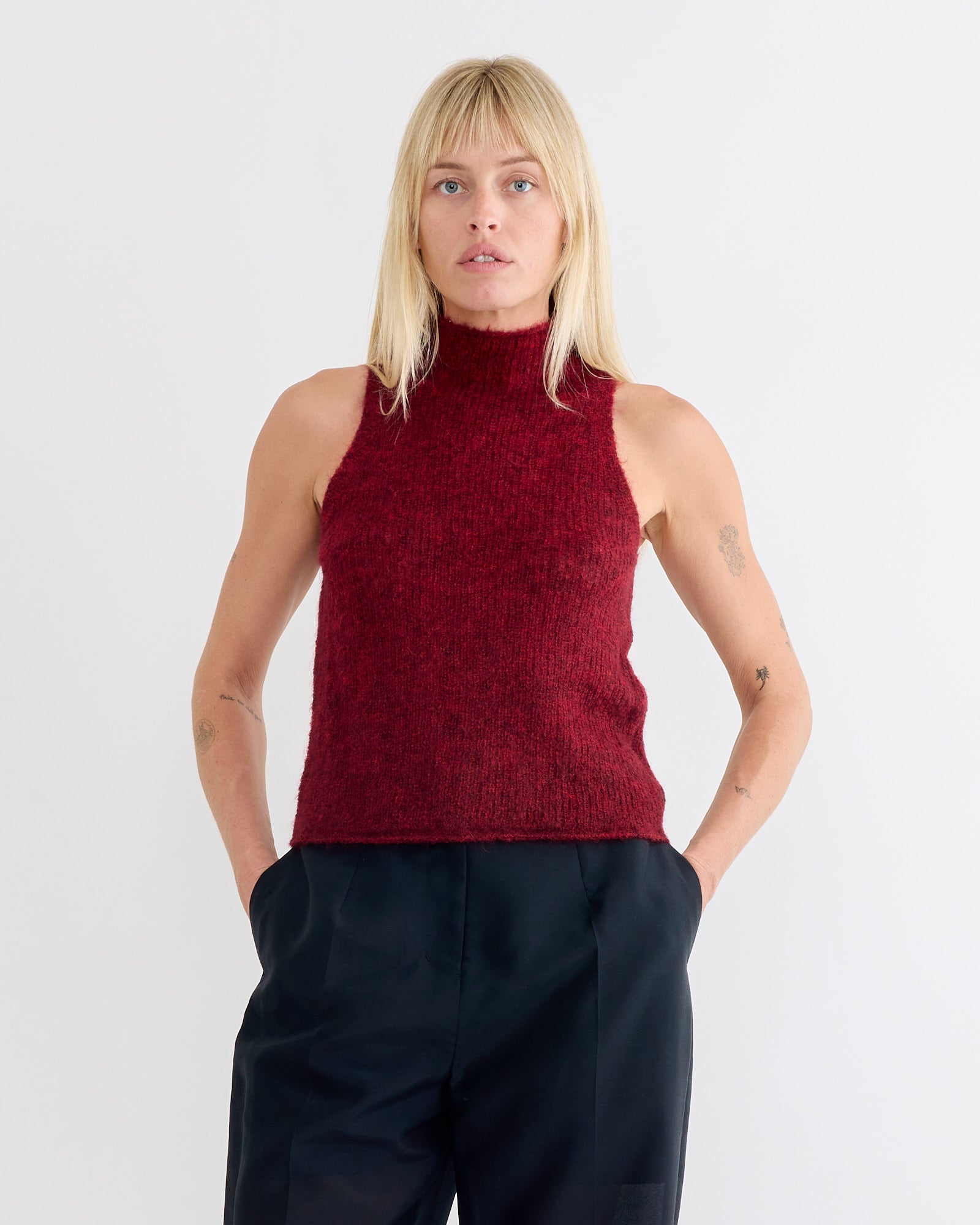 A person with blonde hair wears the Amomento Sheer Sleeveless Top in Red and black pants, standing with hands in pockets against a plain white background.