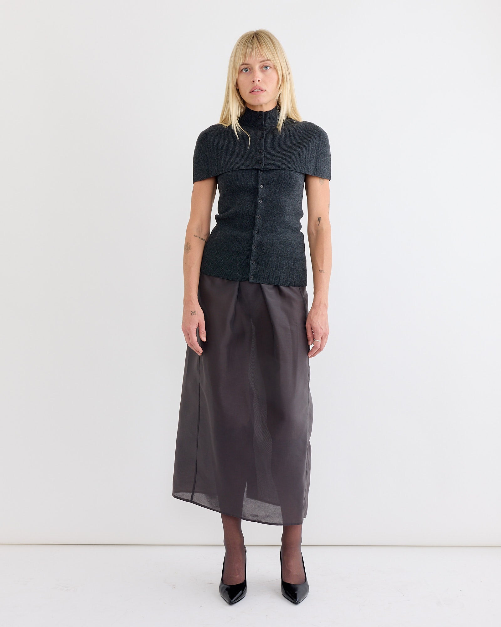 A woman with straight blonde hair wears the Amomento Cape Top & Bandeau Set in Charcoal, paired with a long sheer black skirt and black heels, standing against a plain white wall.