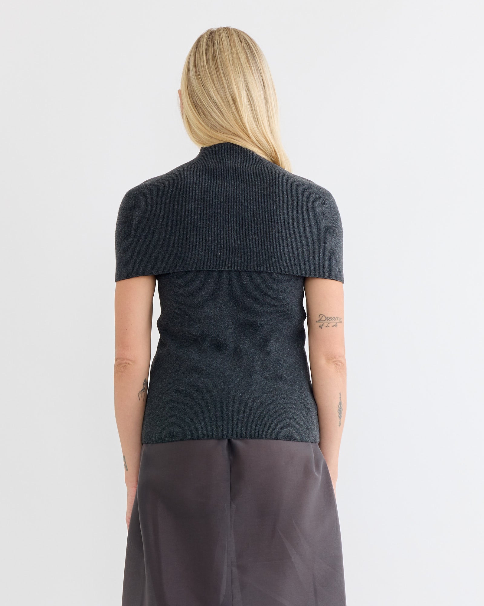 A woman with long blonde hair stands facing away in the Amomento Cape Top & Bandeau Set in Charcoal. She pairs it with a gray skirt, arms relaxed at her sides showing small tattoos, against a plain white background.