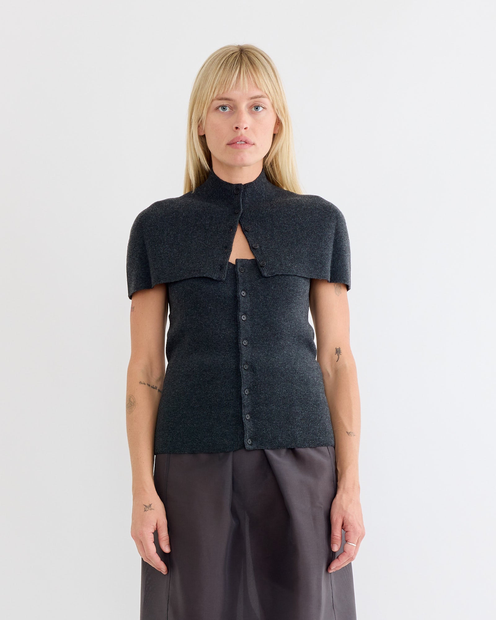 A woman with straight blonde hair models the Amomento Cape Top & Bandeau Set in Charcoal, featuring a high-collar, short-sleeve knit top and matching pants. She stands against a light background, gazing neutrally at the camera.