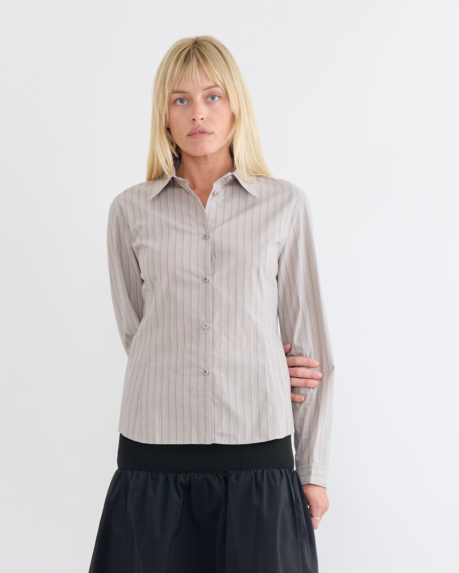 A woman with straight blonde hair wears the Amomento Stripe Shirt in Grey—a striped cotton shirt with a slimming silhouette—and a black skirt, standing against a plain white background with a neutral expression.