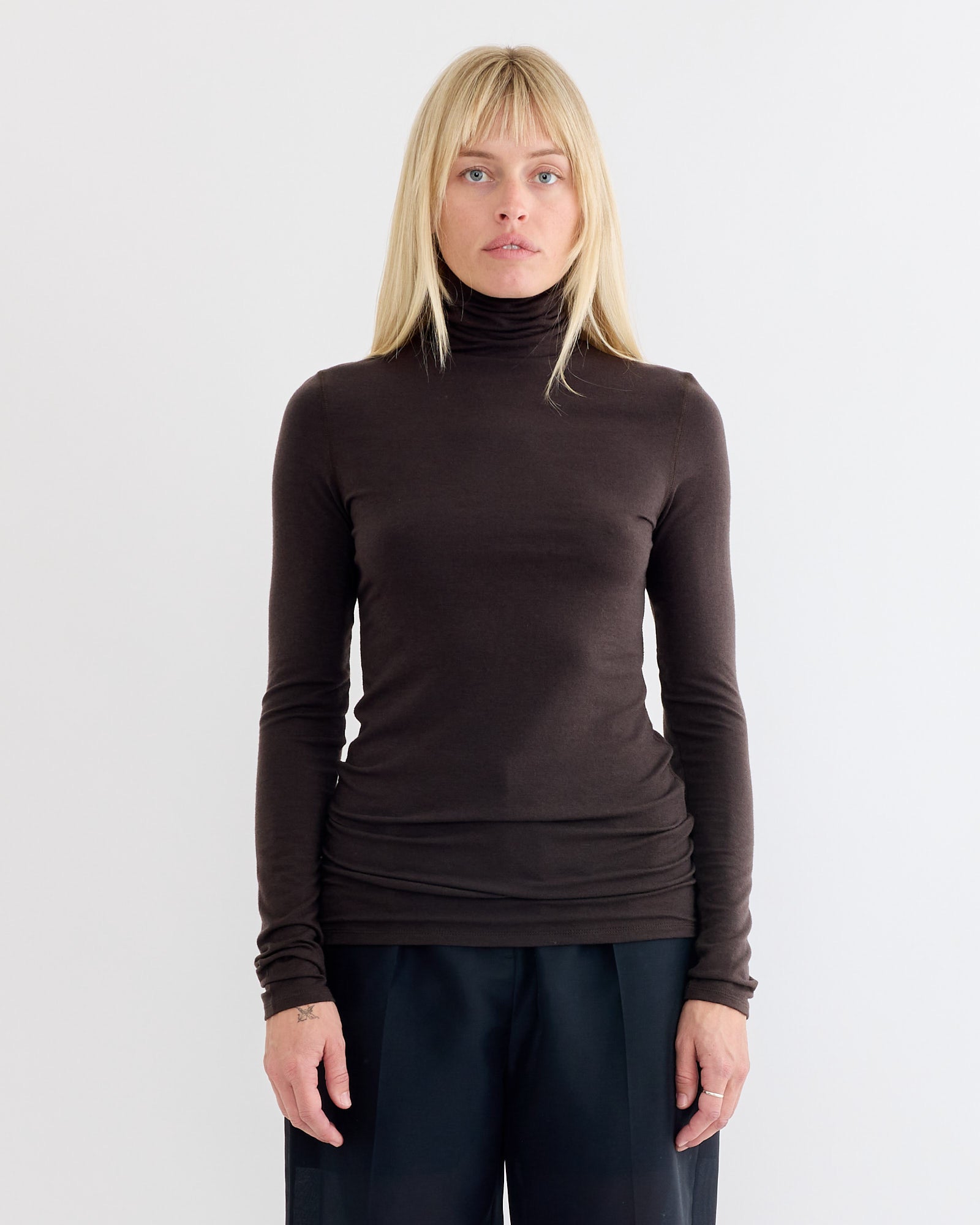 A woman with straight blonde hair wears the Amomento Turtle T-Shirt in Brown as a lightweight winter layer with black pants, standing against a plain light background and looking directly at the camera with a neutral expression.