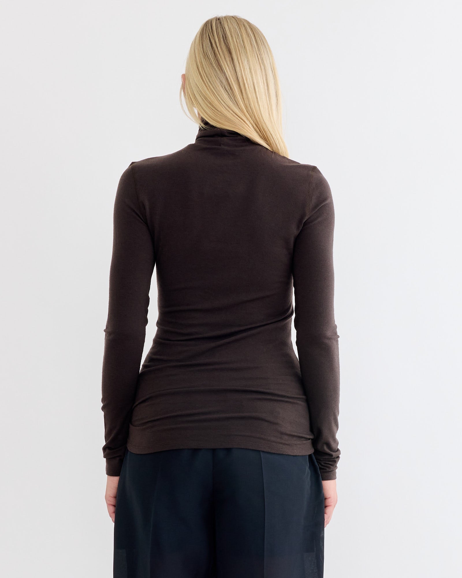 A person with long blonde hair, seen from behind, wears the fitted Amomento Turtle T-Shirt in Brown—an ideal lightweight winter layer—with loose, dark navy pants against a plain, light background.