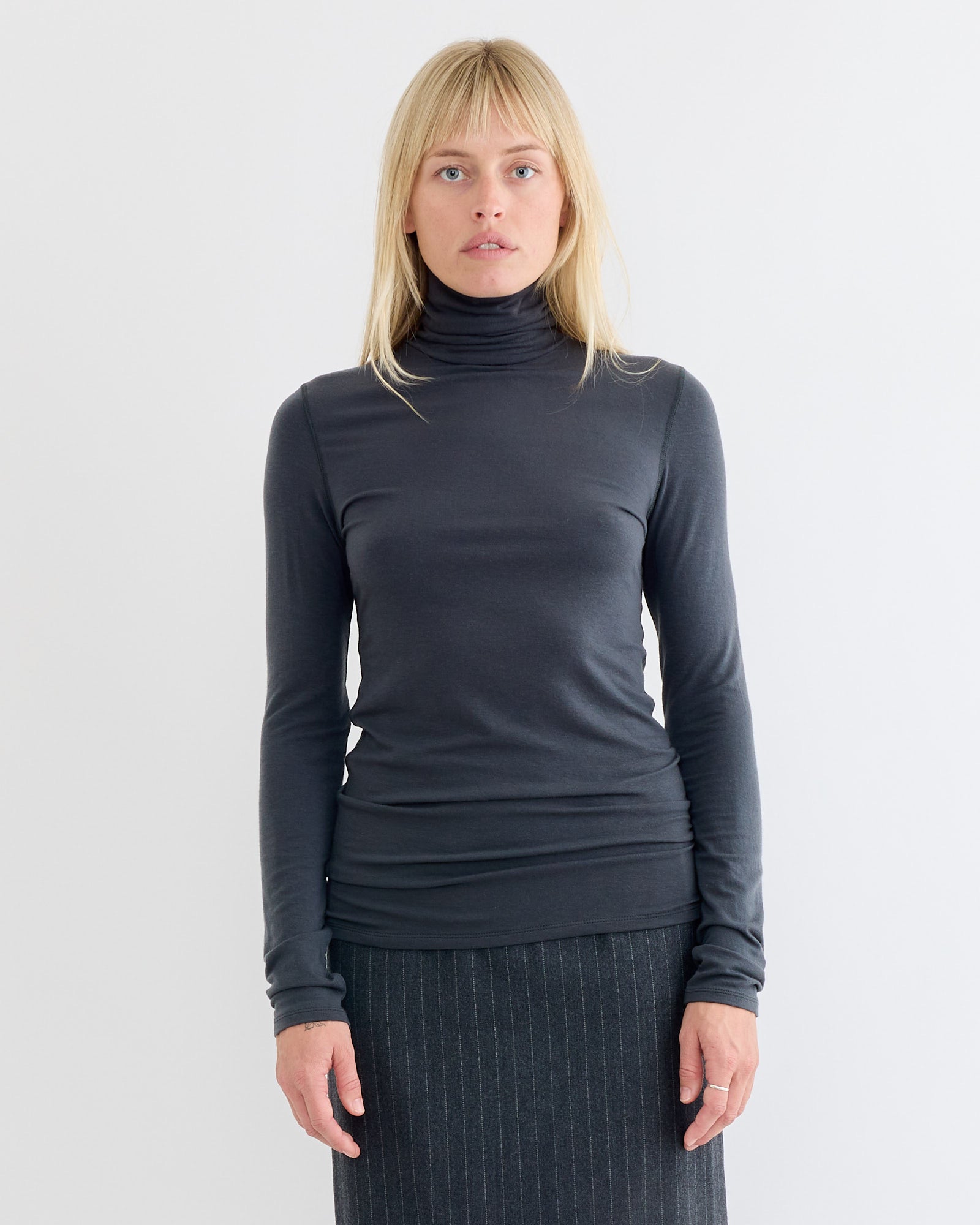 A woman with straight blonde hair models the Amomento Turtle T-Shirt in Charcoal, a lightweight winter layer, paired with a gray pinstripe skirt. She stands against a plain white background, gazing directly at the camera with a neutral expression.