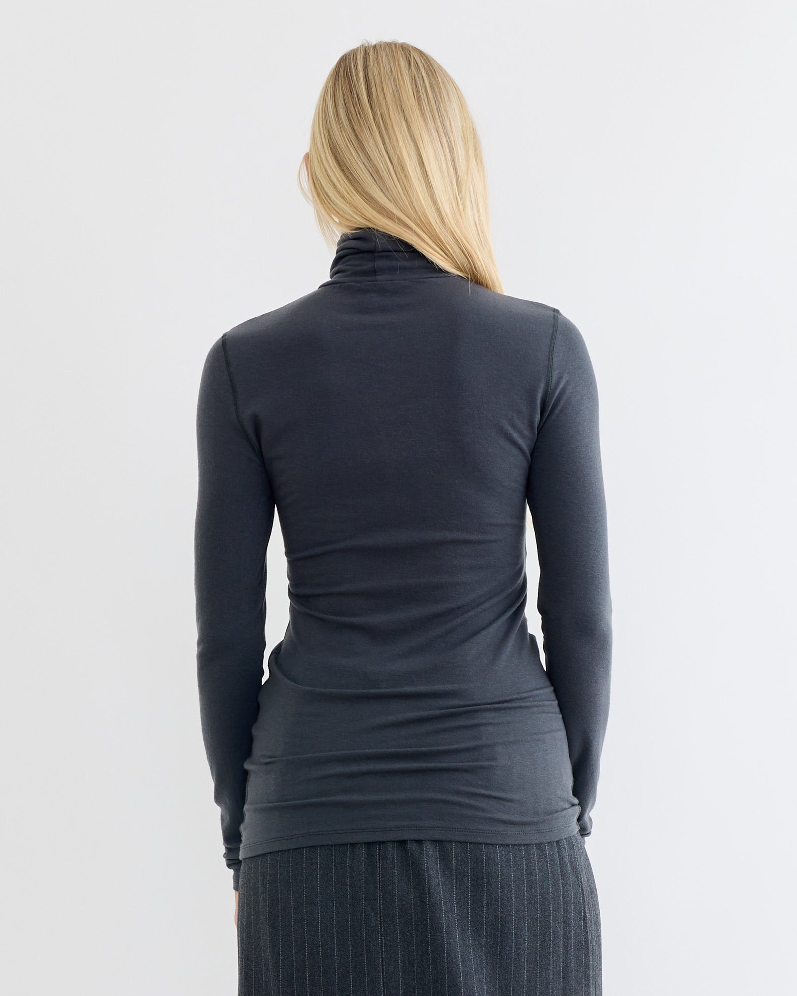 A person with long blonde hair, seen from behind, wears the Amomento Turtle T-Shirt in Charcoal—a fitted, lightweight winter piece—with a pinstriped skirt, set against a plain, light background.