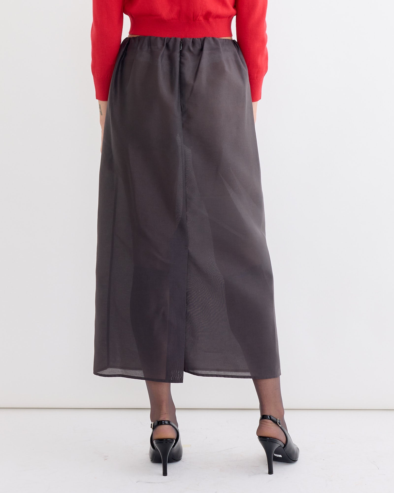 A person in the Amomento Organza Layered Skirt in Charcoal, black heels, and a red top stands with their back to the camera against a plain white background.