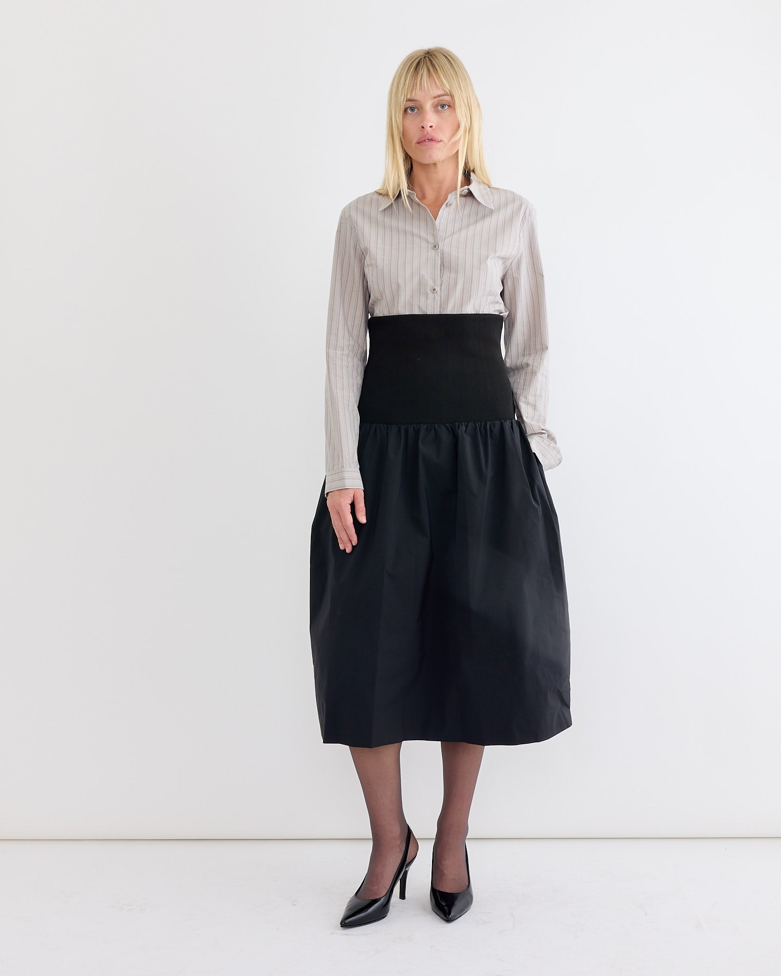 A woman with blonde hair stands against a white wall, wearing the Amomento Stripe Shirt in Grey tucked into a high-waisted black midi skirt. She pairs it with black heels and sheer tights, creating a chic, slimming look as she faces the camera.