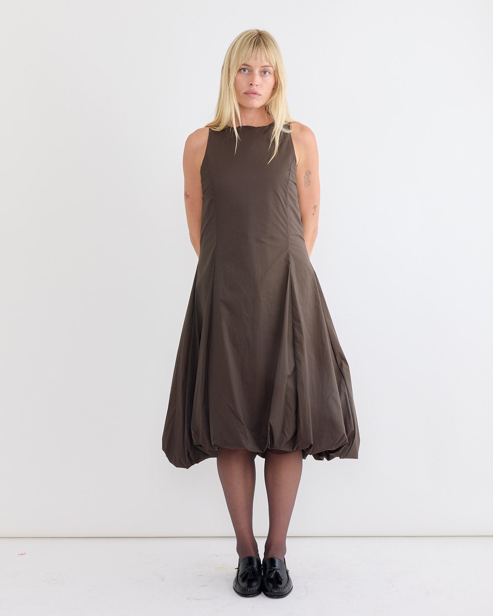A woman with blonde hair stands against a white background, wearing Amomento’s Sheer Padded Volume Dress in Brown, styled with sheer black tights and black shoes. She faces forward with a neutral expression.