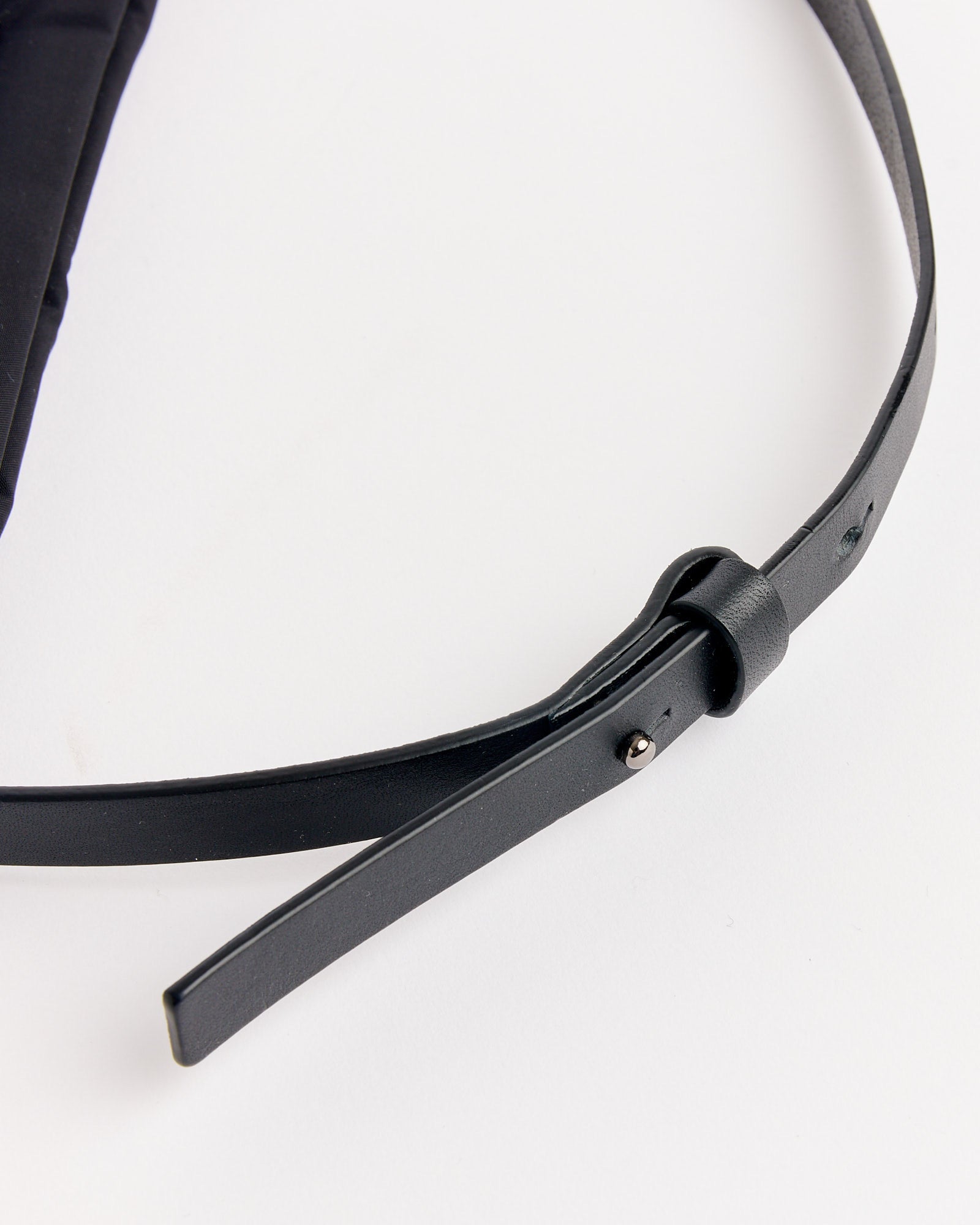 A close-up of the Amomento Mini Padded Bag in Black, featuring leather straps and a simple metal stud fastening, shown on a white surface with the strap partially threaded through its own loop.