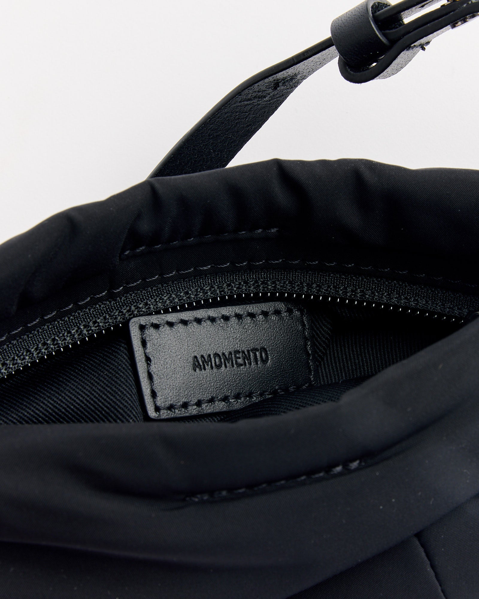 Close-up of the Amomento Mini Padded Bag in Black, featuring a visible zipper, stitched black leather "AMENDMENT" label inside, and partially visible black strap with buckle and leather details at the top.