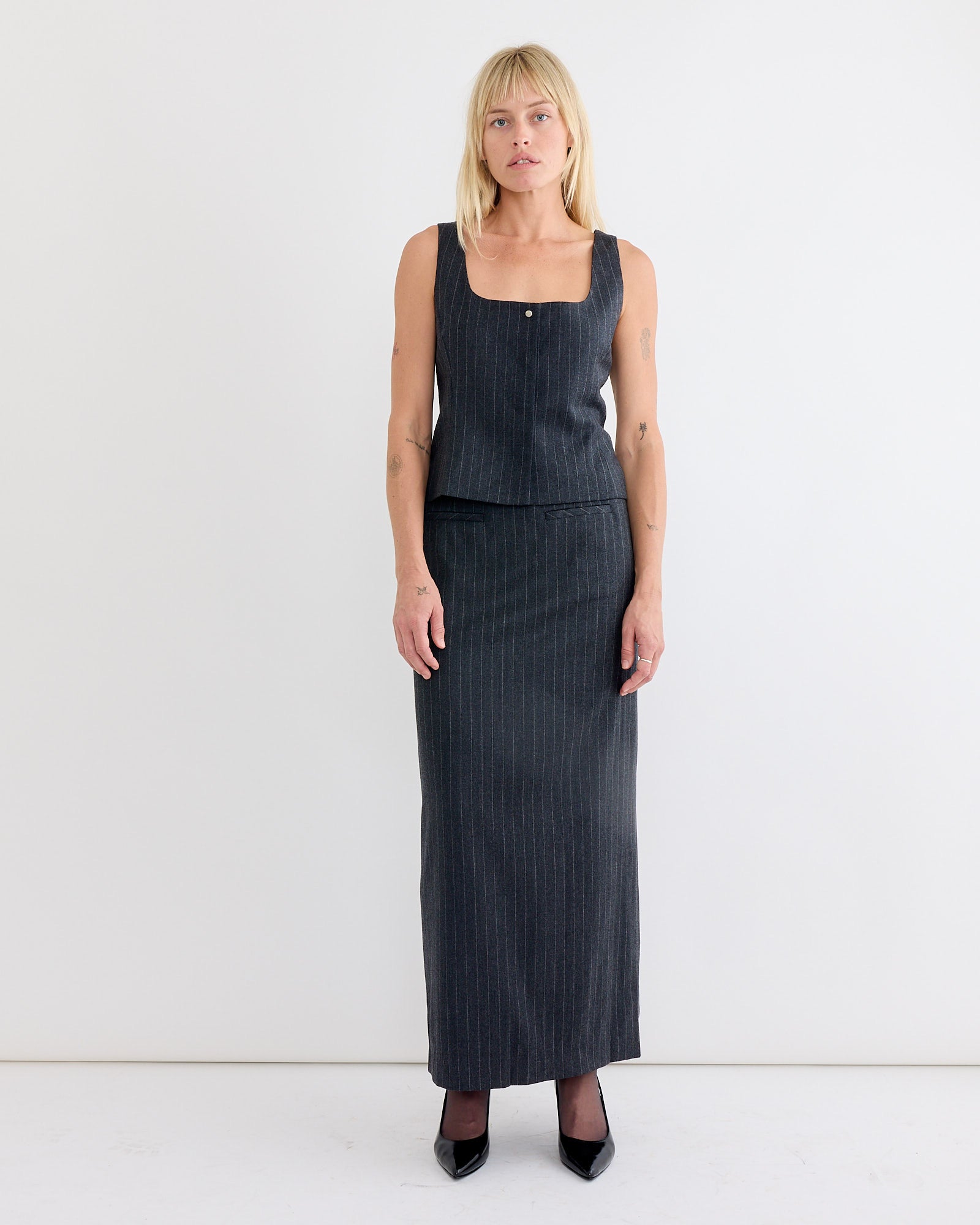 A woman with blonde bangs stands against a white background wearing Amomento's Wool Snap Button Down Vest in Stripe, a dark gray pinstriped sleeveless top with a square neckline, paired with the matching Stripe Maxi Skirt and black heels.