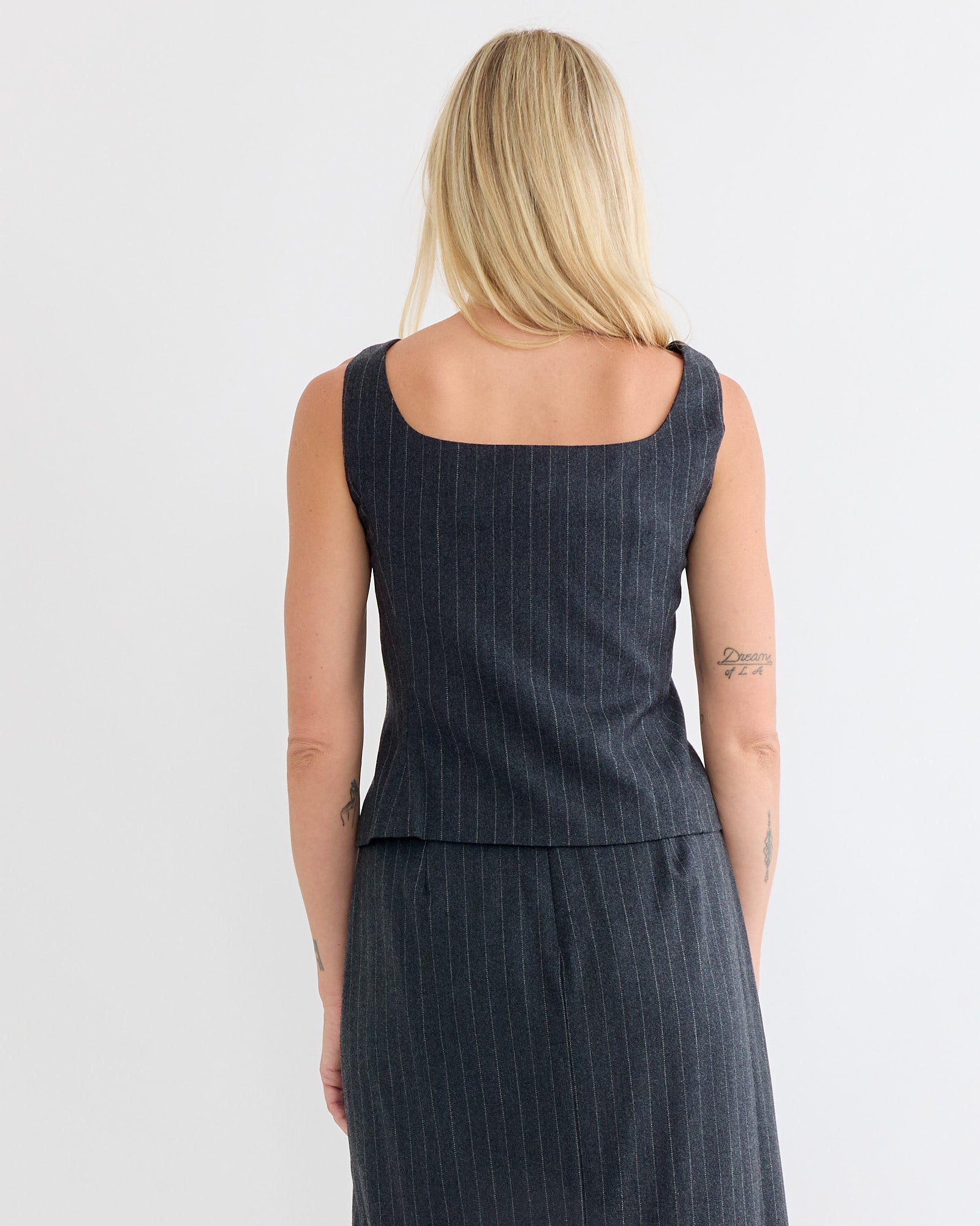 A person with long blonde hair is seen from behind, wearing the Amomento Wool Snap Button Down Vest in Stripe and a matching skirt. The dark gray pinstriped outfit shows their tattooed arms against a plain white background.