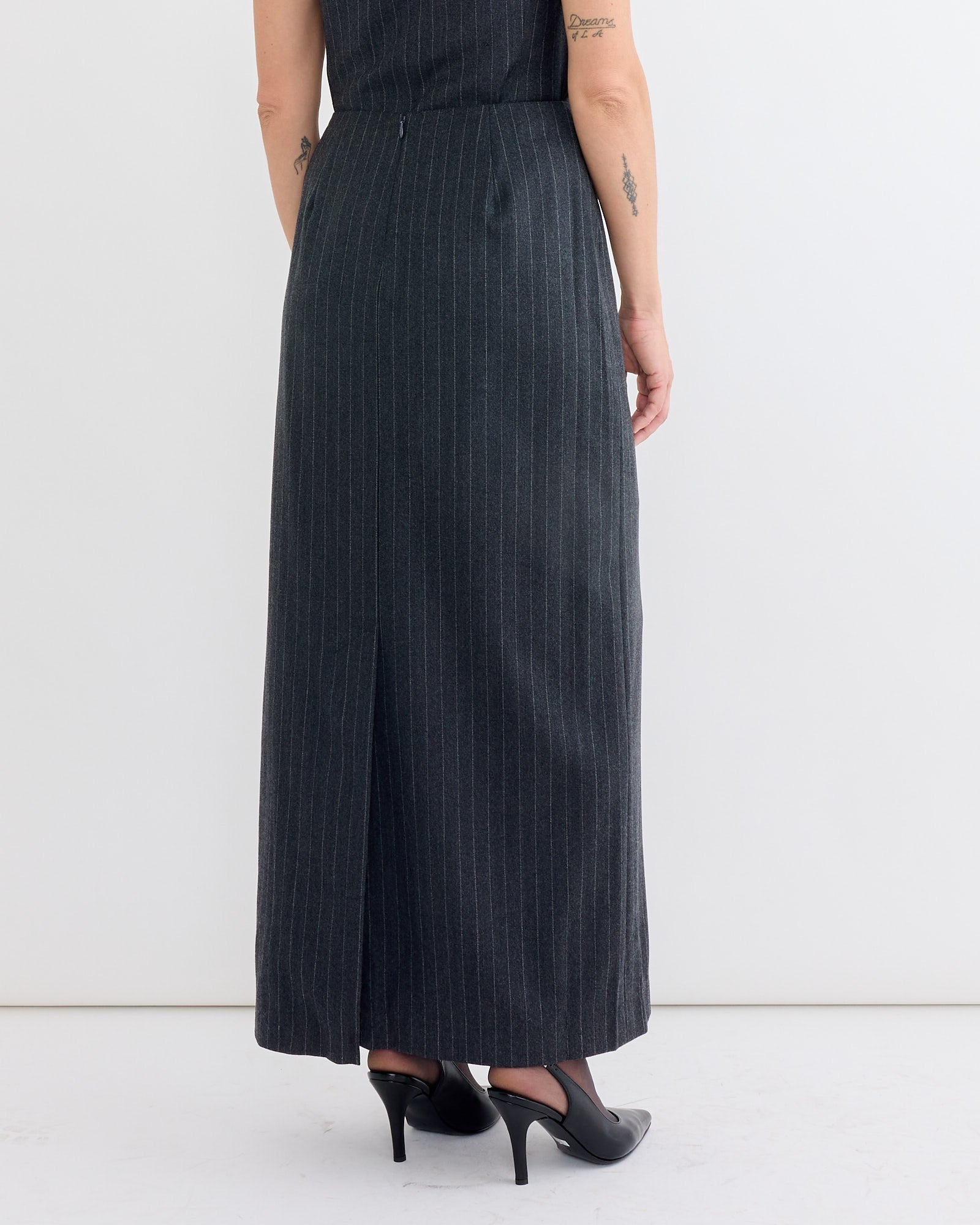 A person stands with their back to the camera, wearing the Amomento Wool Maxi Long Skirt in Stripe and black heels. Their arms hang relaxed at their sides, showing small tattoos, against a plain white background.