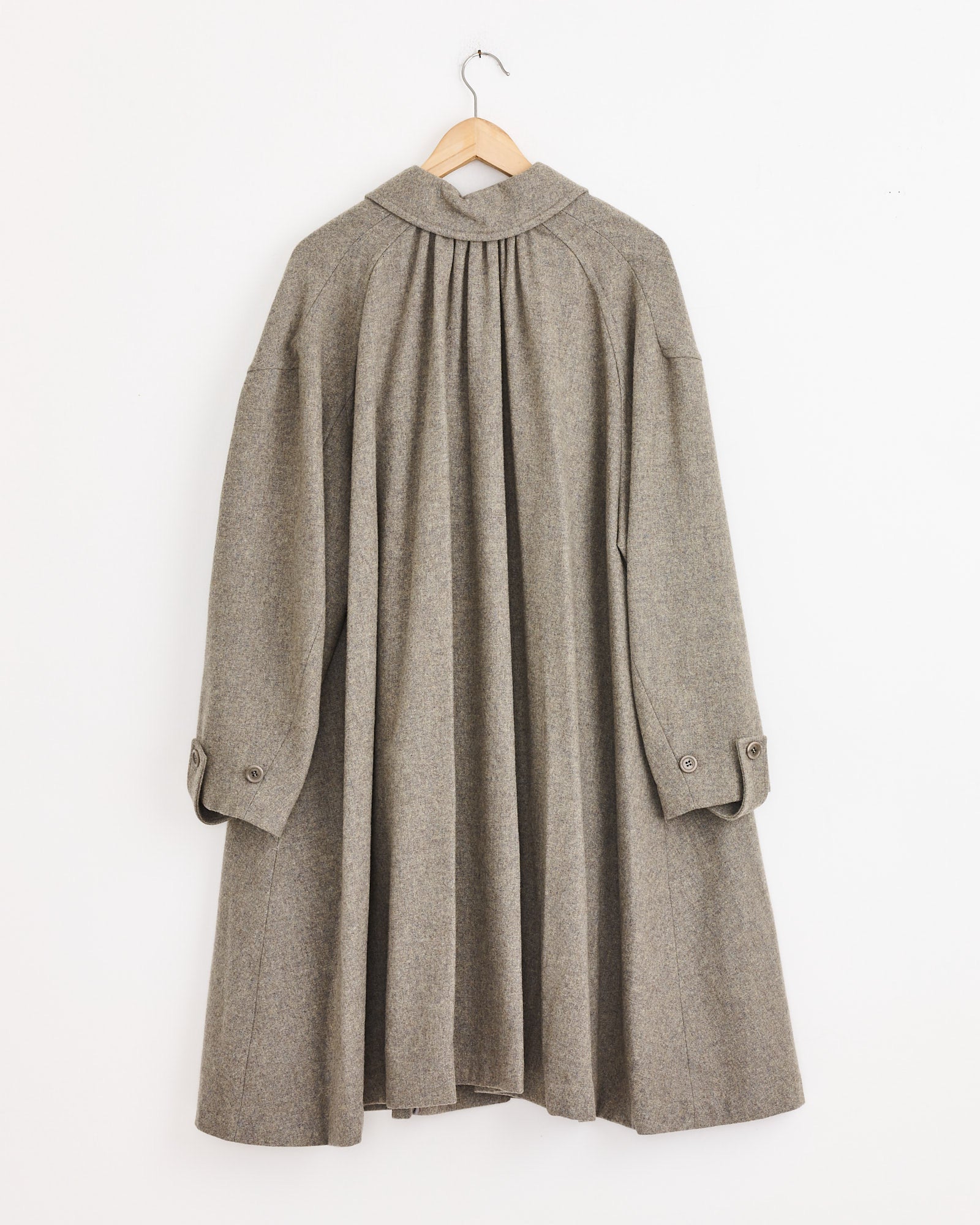 The Amomento Shirring Coat in Grey Blue, featuring wide sleeves, buttoned cuffs, and pleated details, hangs on a wooden hanger against a plain white wall. The back view highlights its subtle flare silhouette.