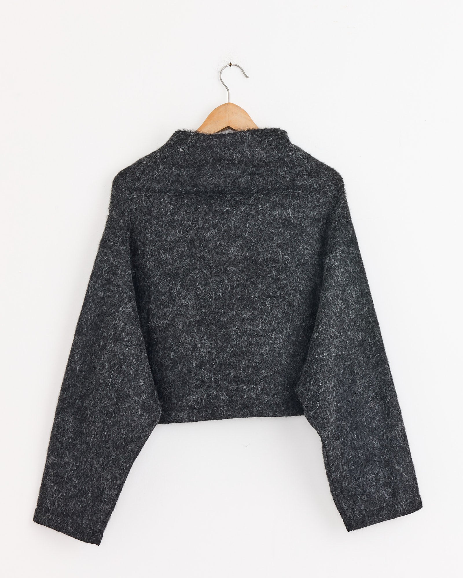 The Amomento Alpaca Top in Charcoal is a lightweight, long-sleeved, cropped sweater made from fuzzy alpaca knit. Shown from the back, it hangs on a wooden hanger against a plain white background.