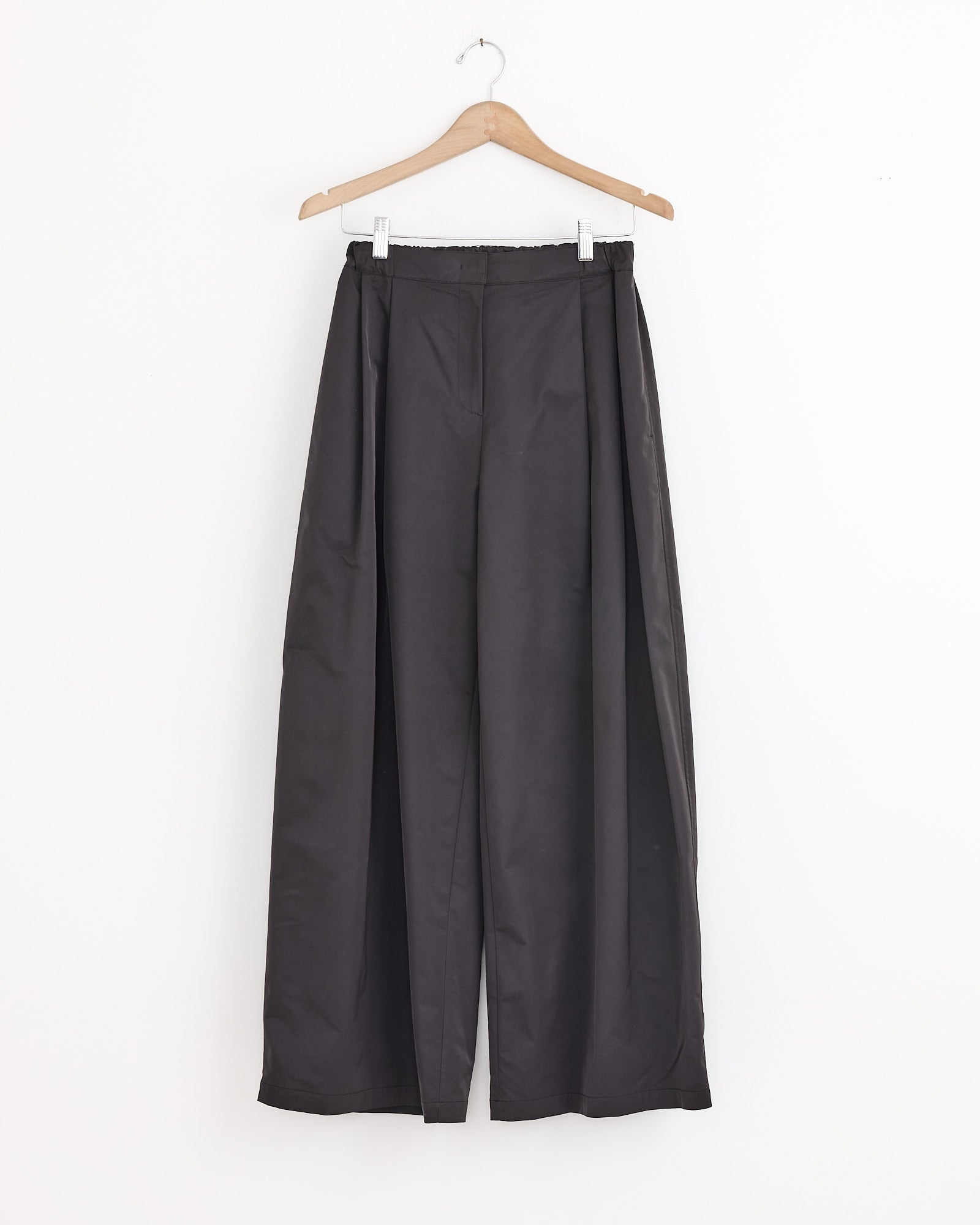 The Amomento Two Tuck Balloon Pant in Charcoal hangs on a wooden hanger against a white background, featuring a wide-leg silhouette, cozy brushed lining, elastic waistband, and minimalist design.
