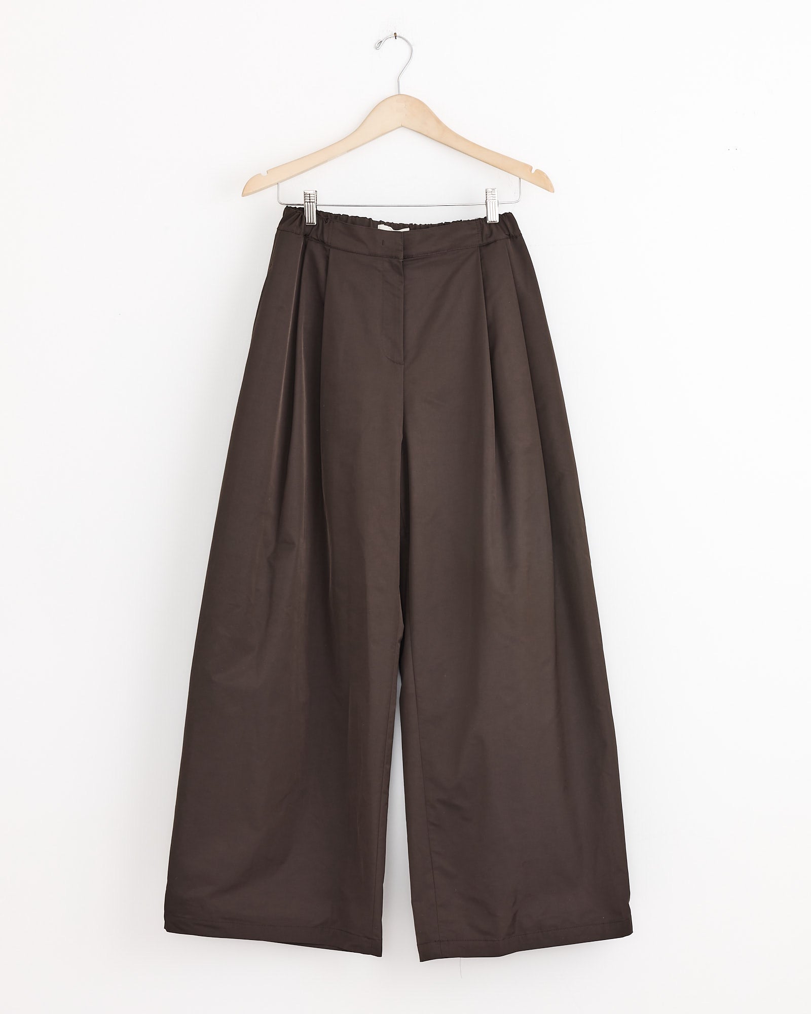 The Amomento Two Tuck Balloon Pant in Dark Brown features a high waist, pleats, and a wide-leg silhouette. Shown hanging on a wooden hanger against a plain white background.