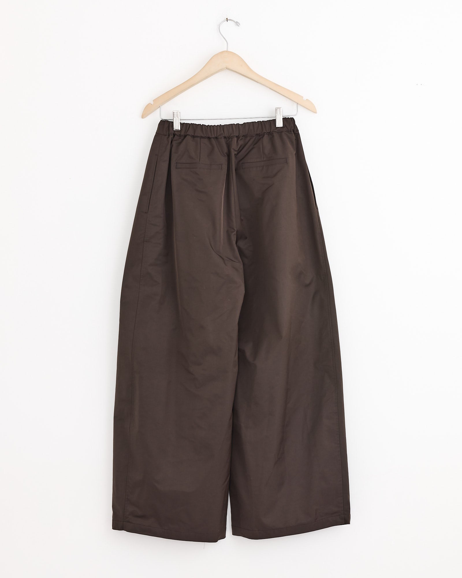 The Amomento Two Tuck Balloon Pant in Dark Brown features a wide-leg silhouette and an elastic waistband, displayed on a wooden hanger against a white background.
