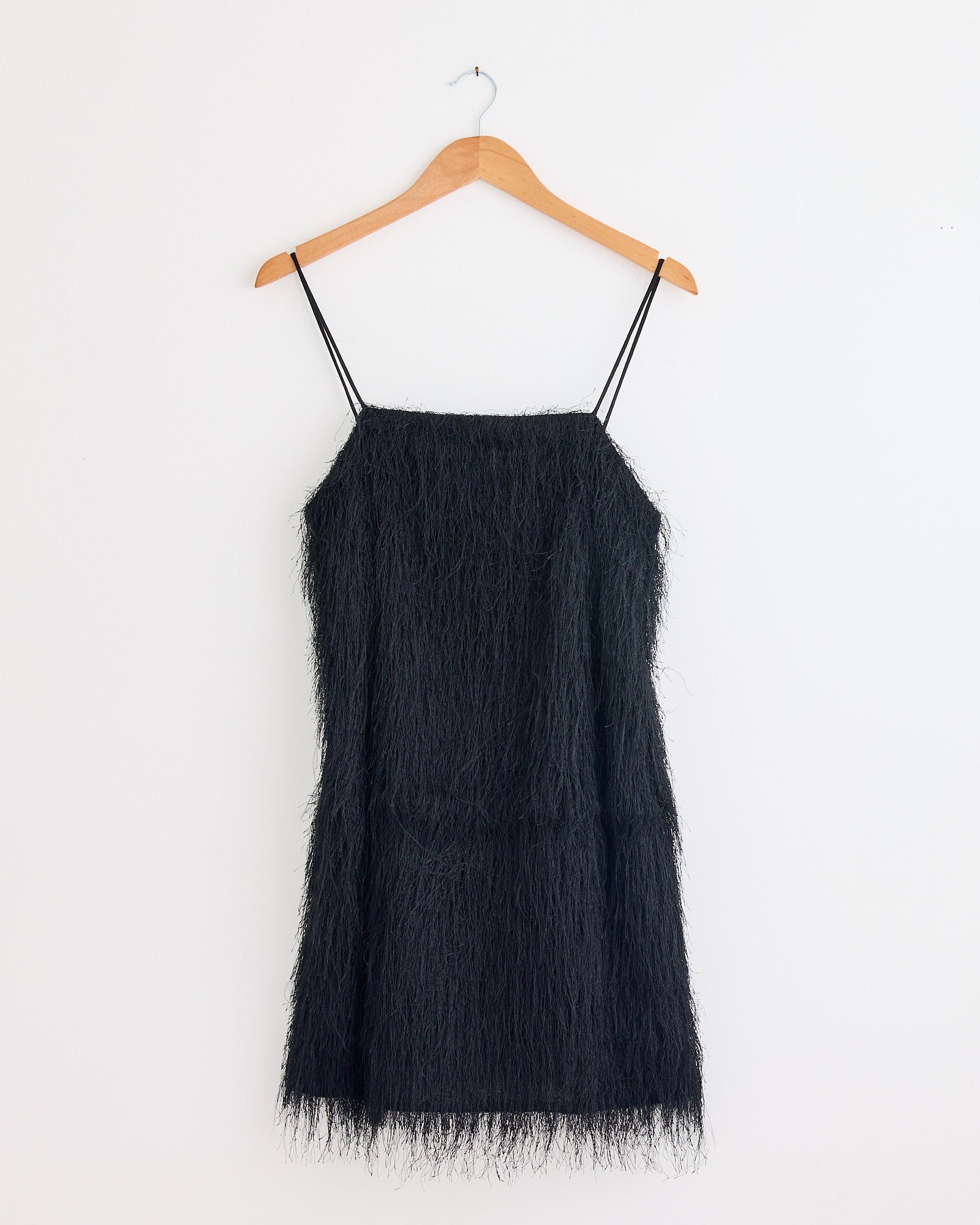 The Ledo Dress in Black by By Malene Birger, featuring a fringe mini silhouette and thin spaghetti straps, hangs on a wooden hanger against a white wall—ideal for chic eveningwear.