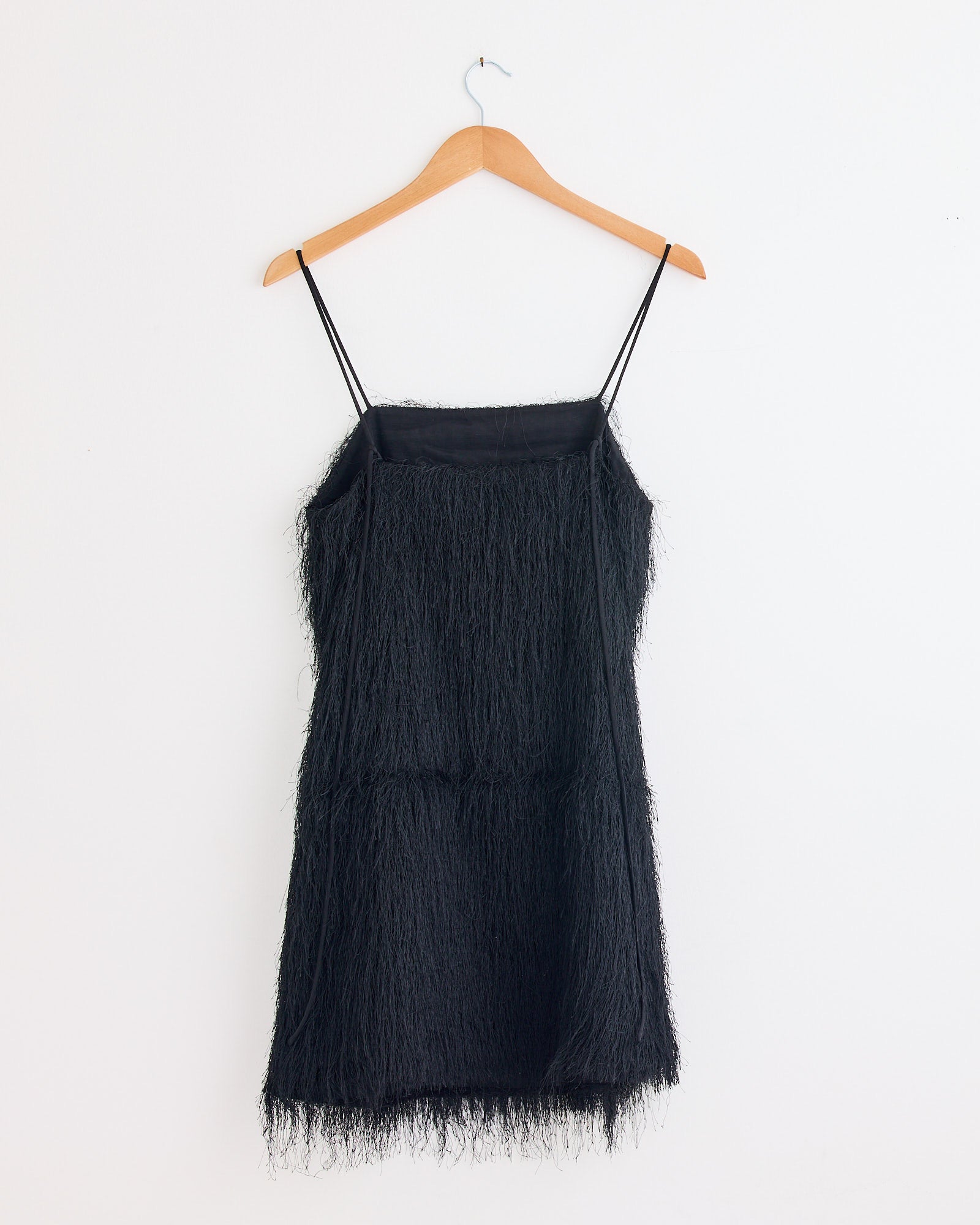 The Ledo Dress in Black by By Malene Birger—a black, sleeveless mini dress with spaghetti straps and textured fringe—hangs on a wooden hanger against a plain white wall, offering an elegant eveningwear option.