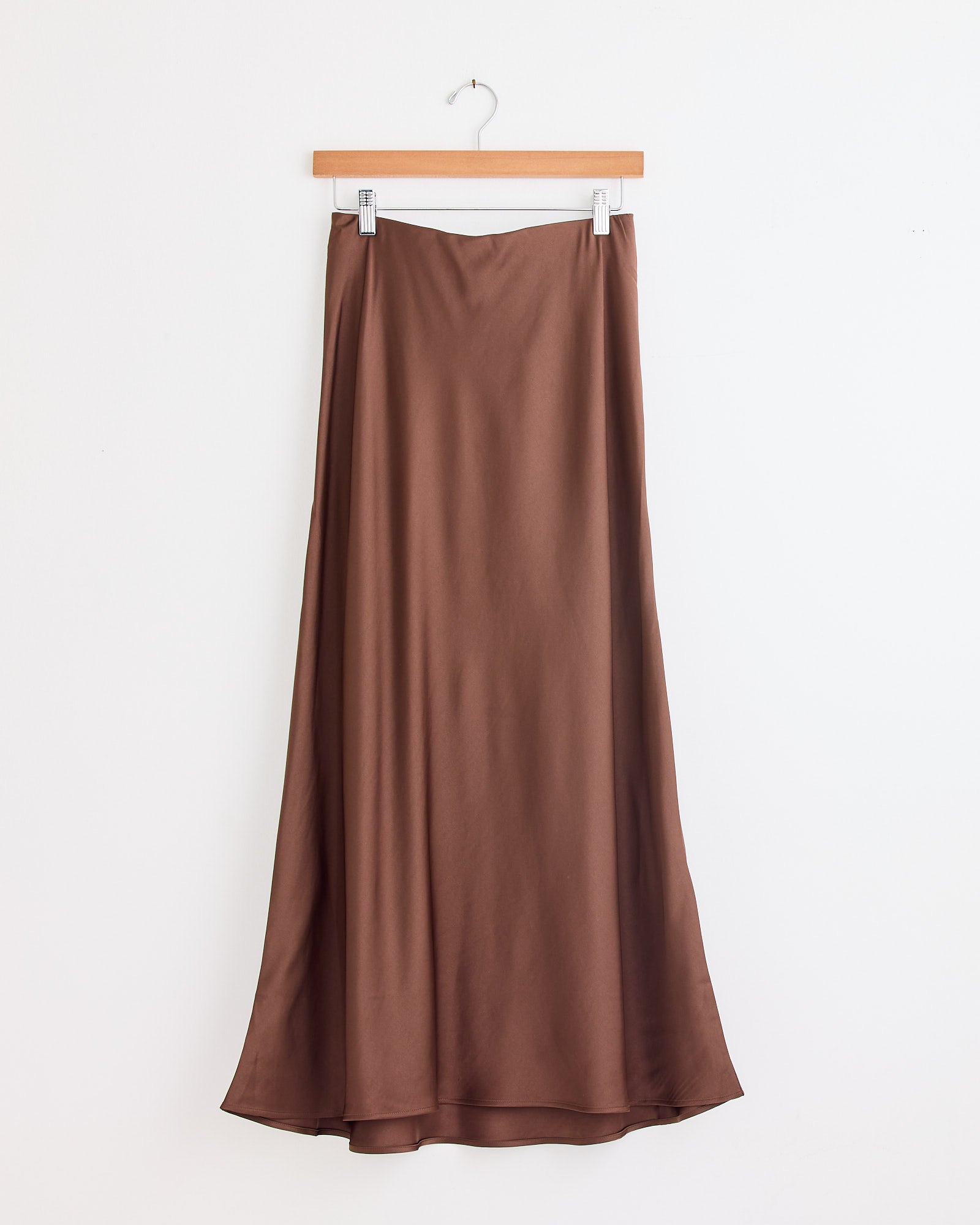 The Boshan Skirt in Carafe by By Malene Birger, a midi-length A-line skirt with a smooth satin finish and subtle flare, hangs on a wooden hanger against a white wall.