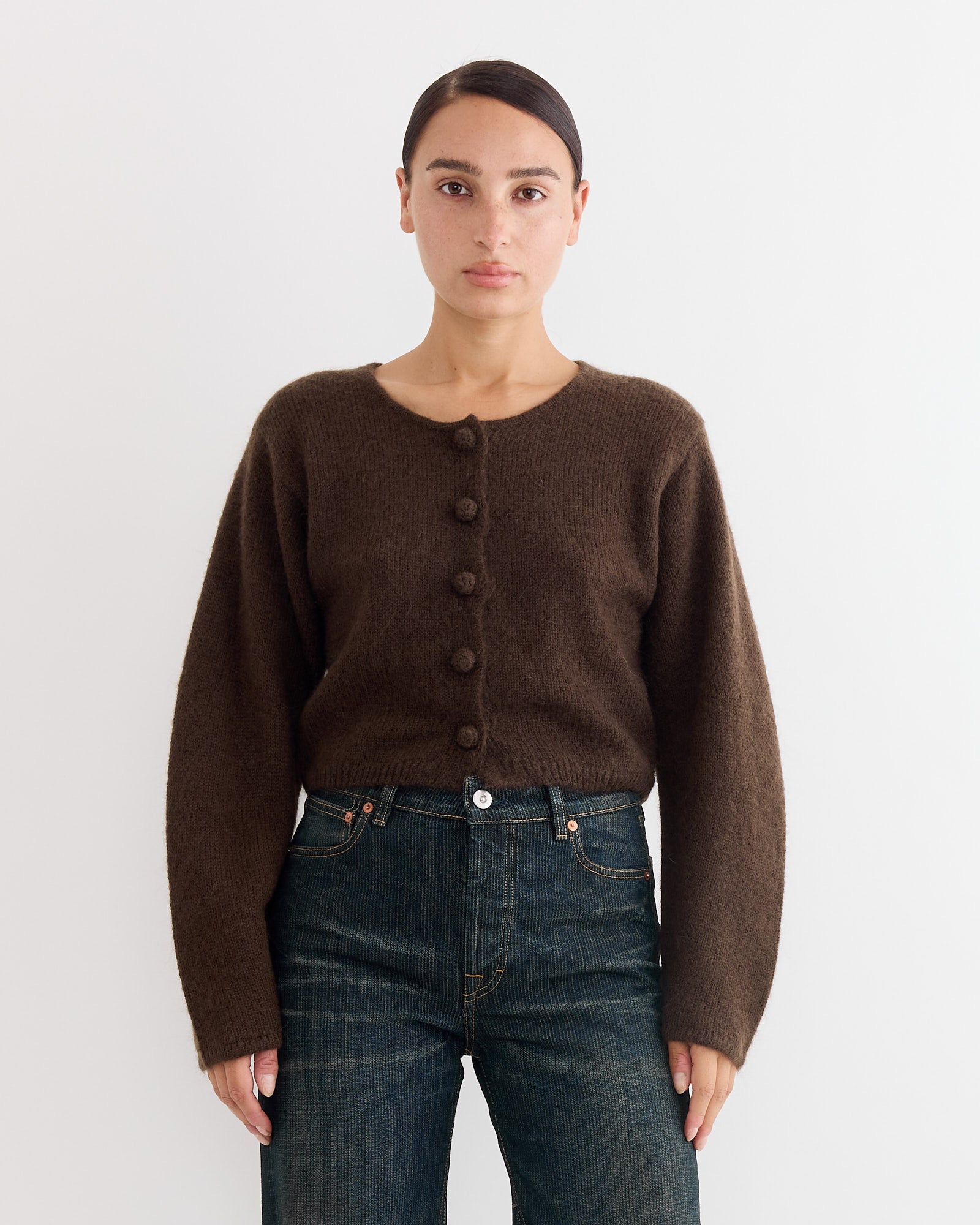 A person with dark hair pulled back wears a By Malene Birger Zharea Cardigan in Brown Mahogany and dark blue jeans, standing against a plain white background.