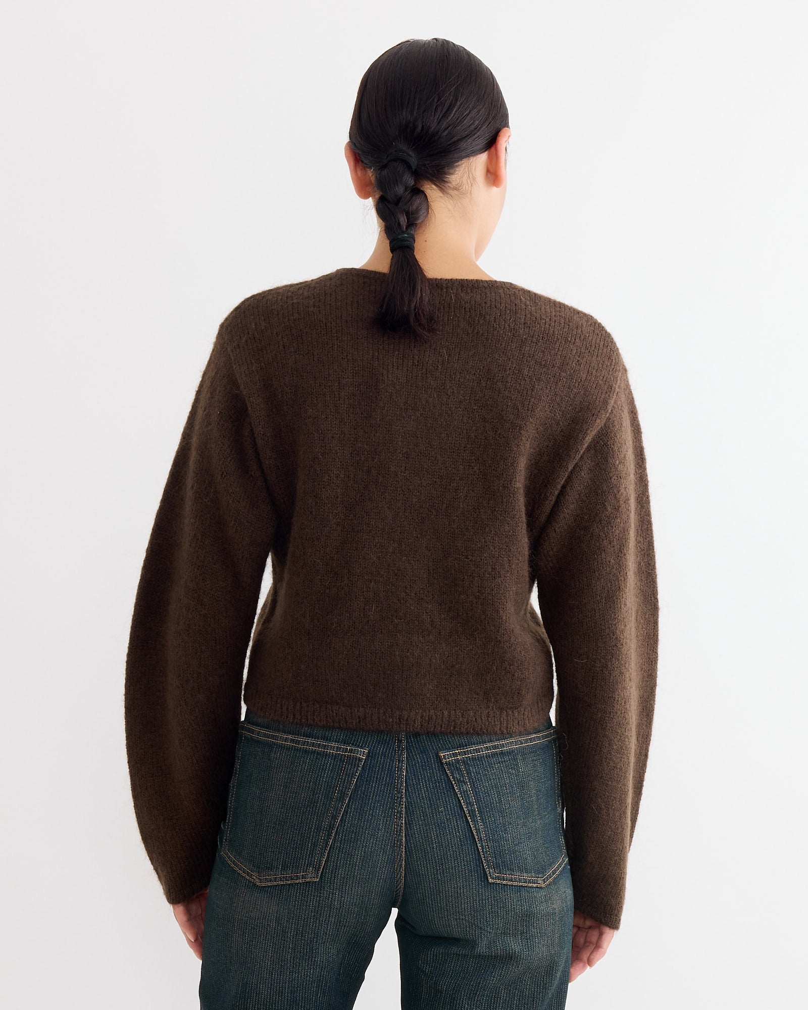 A person with dark braided hair is seen from behind, wearing a By Malene Birger Zharea Cardigan in Brown Mahogany and blue cropped trousers against a plain white background.