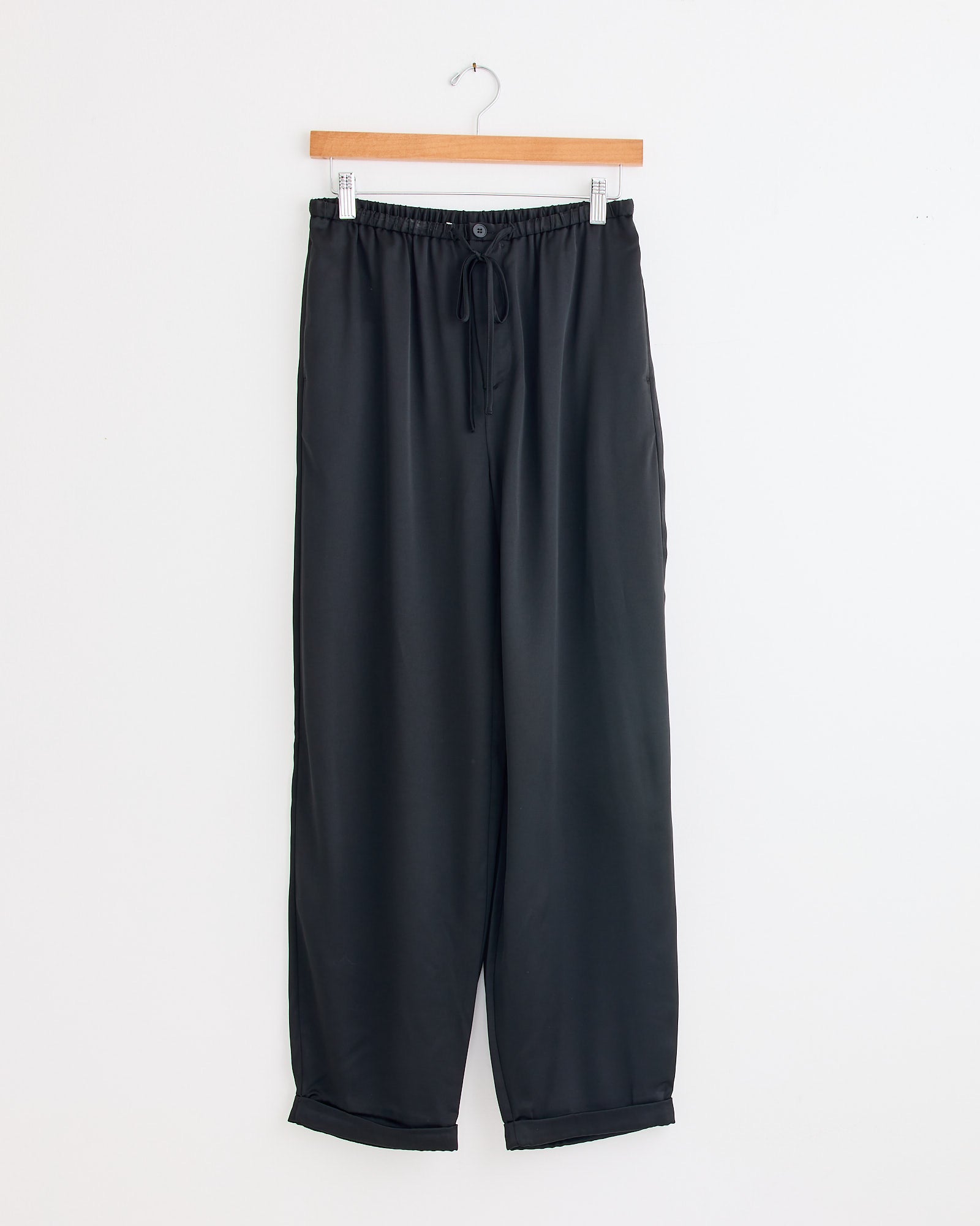 The Joannie Satin Pant in Black by By Malene Birger hangs on a wooden hanger against a white wall. These wide-leg satin pants offer a relaxed fit, elastic waistband with button, and a subtle, elegant shine.