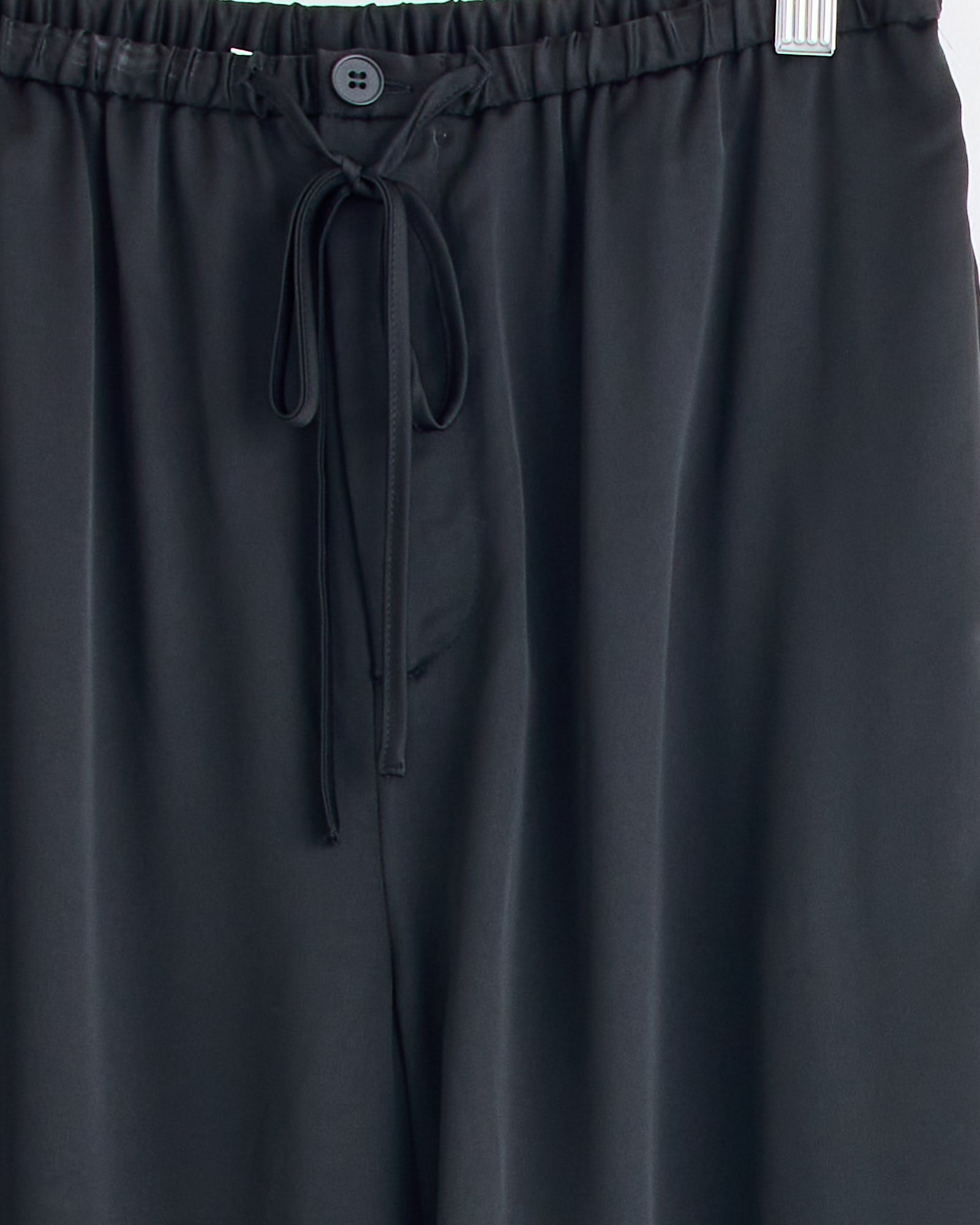 Close-up of the Joannie Satin Pant in Black by By Malene Birger, hanging on a hanger. Features an elastic waistband with button and tie, wide-leg fit, and smooth, slightly shiny satin fabric.