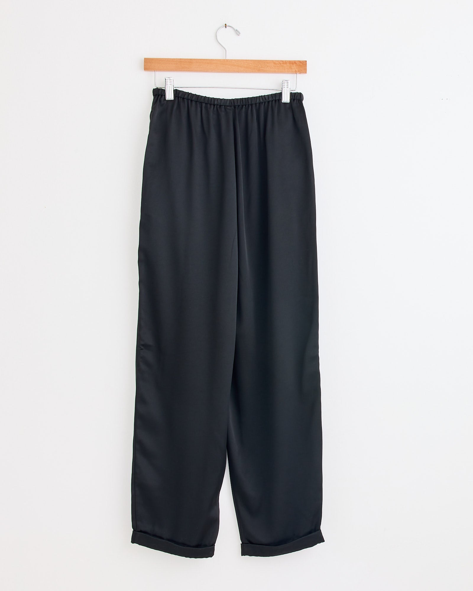 The Joannie Satin Pant in Black by By Malene Birger, featuring a loose wide-leg fit and elastic waistband, hangs on a wooden hanger against a plain white wall.