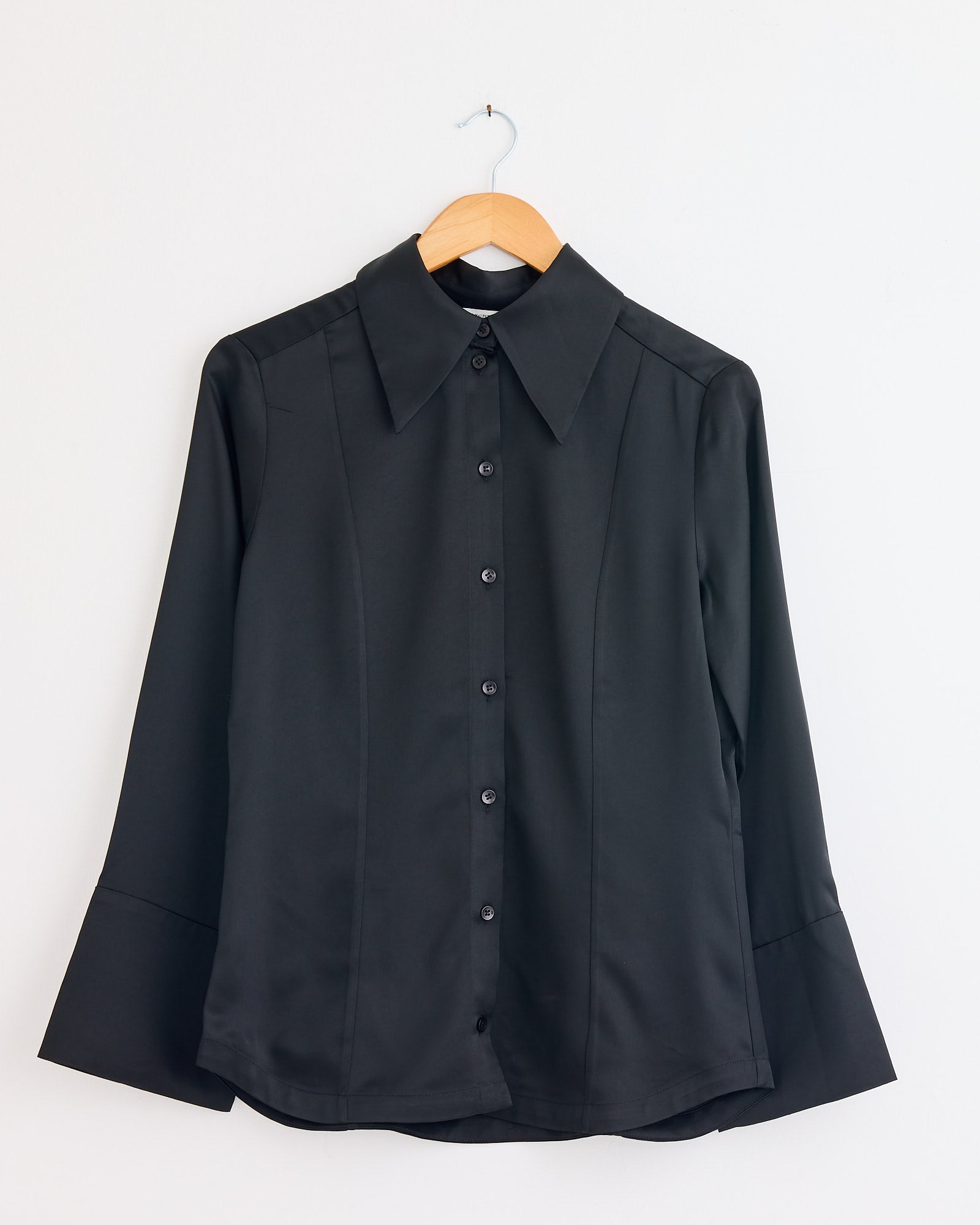 The Rosiannas Satin Blouse in Black by By Malene Birger, with long sleeves, a wide collar, and an elegant fit, hangs on a wooden hanger against a plain white wall.