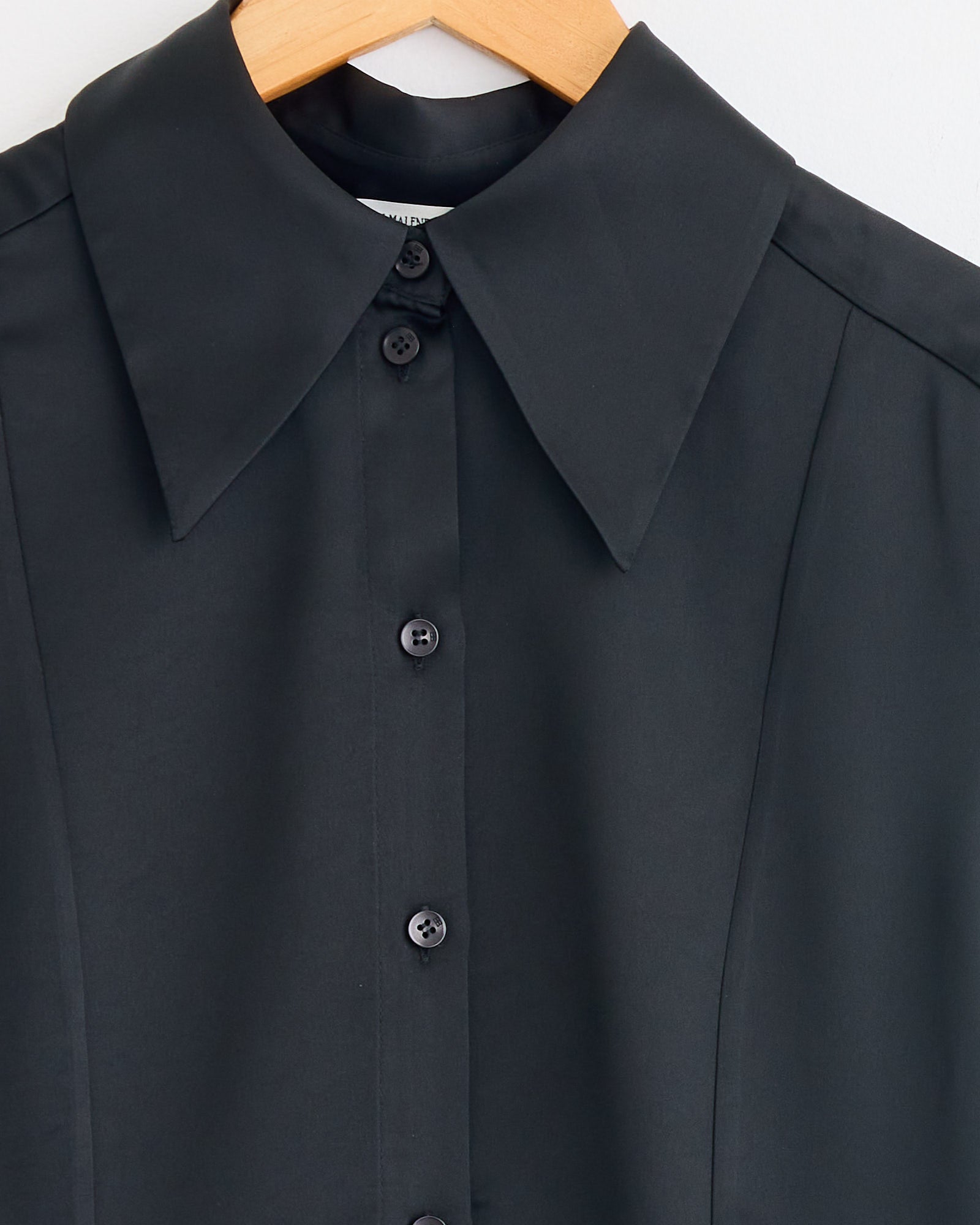 Close-up of the By Malene Birger Rosiannas Satin Blouse in Black, featuring a wide, pointed collar and elegant fit, displayed on a wooden hanger against a white background.