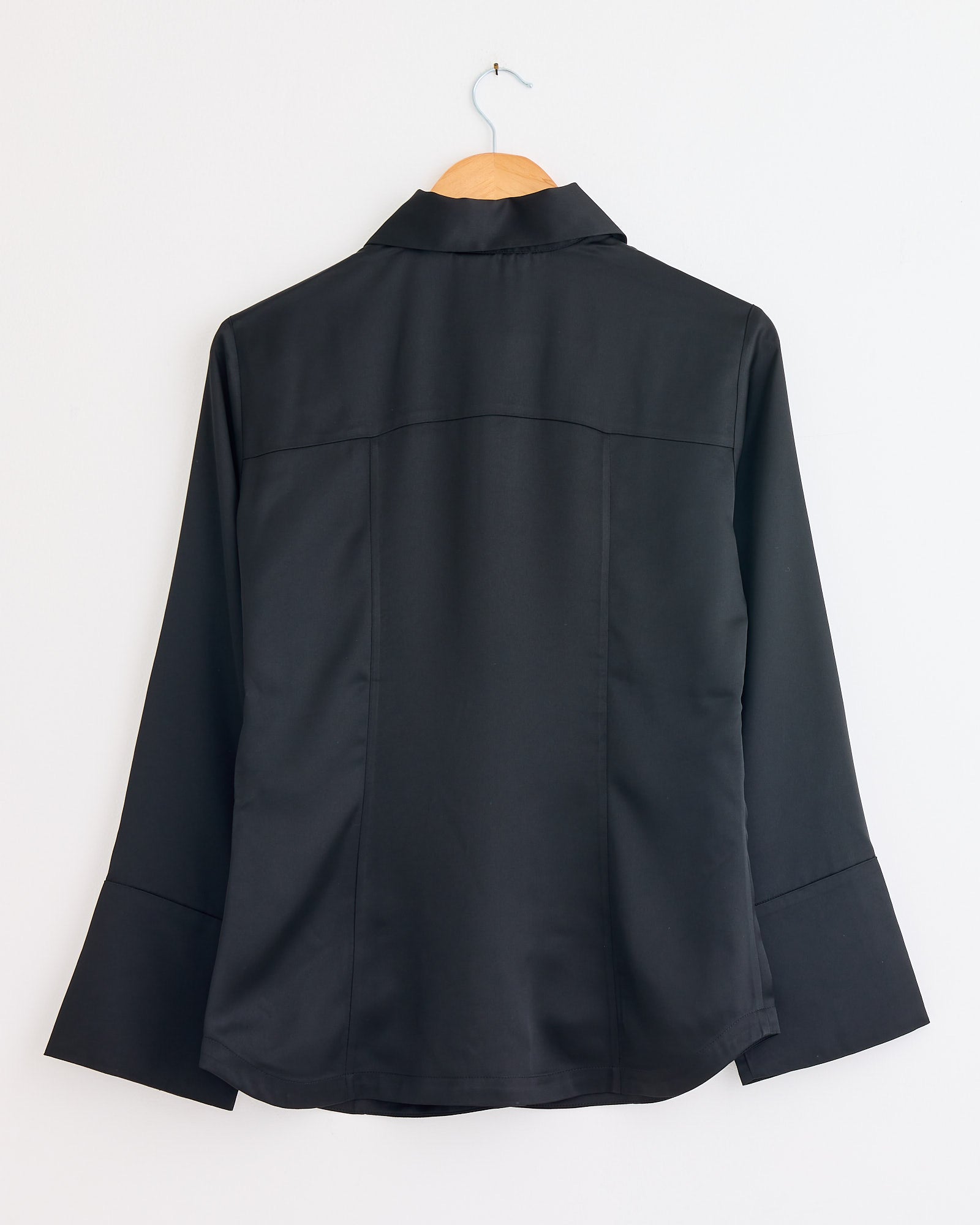 The Rosiannas Satin Blouse in Black by By Malene Birger hangs on a wooden hanger against a white wall, displayed from the back to highlight its elegant fit and long sleeves.