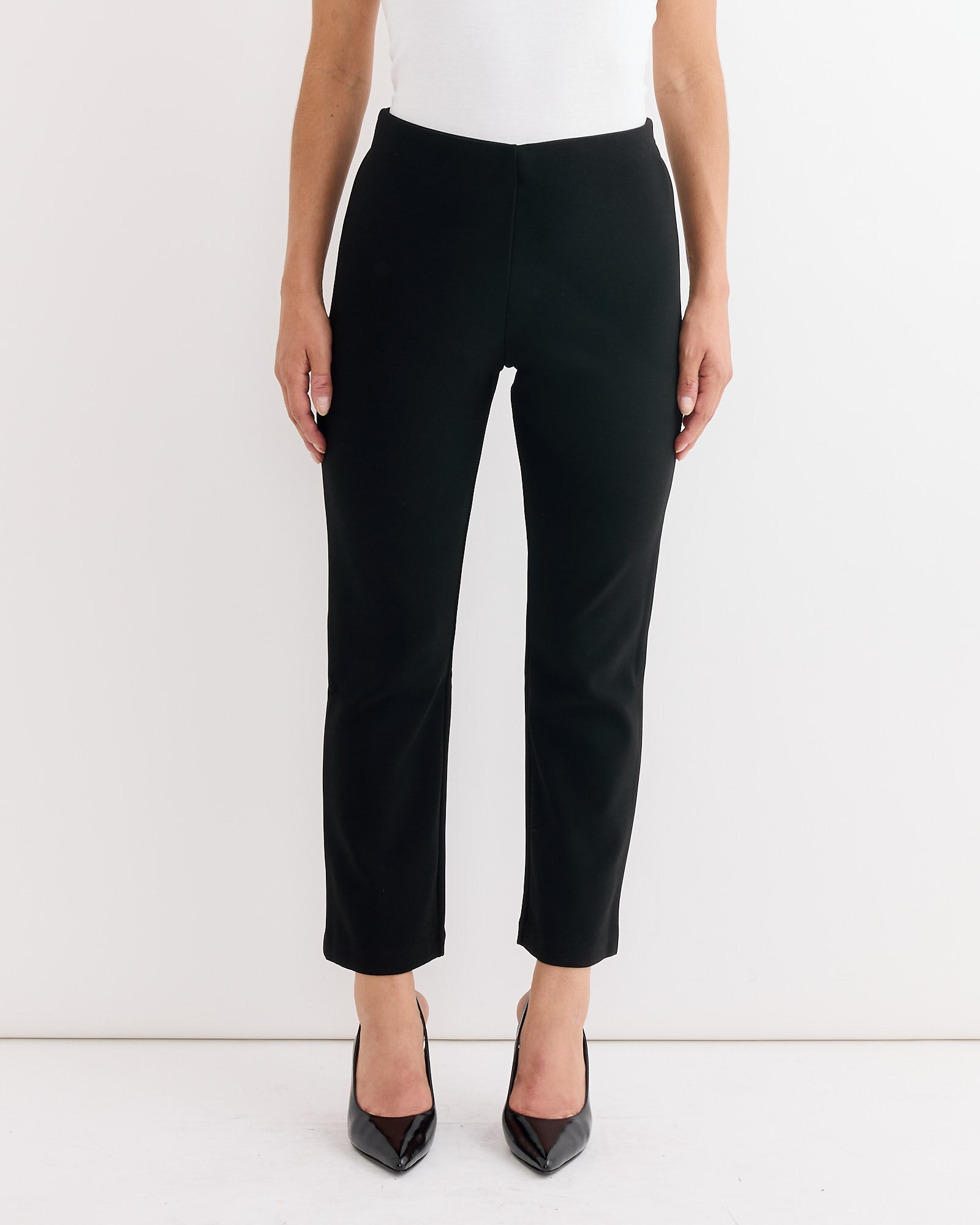 A person models the Vilea Cropped Trouser in Black by By Malene Birger with black pointed-toe heels, cropped from shoulders down against a white background. For sizing inquiries, please contact us via online chat.