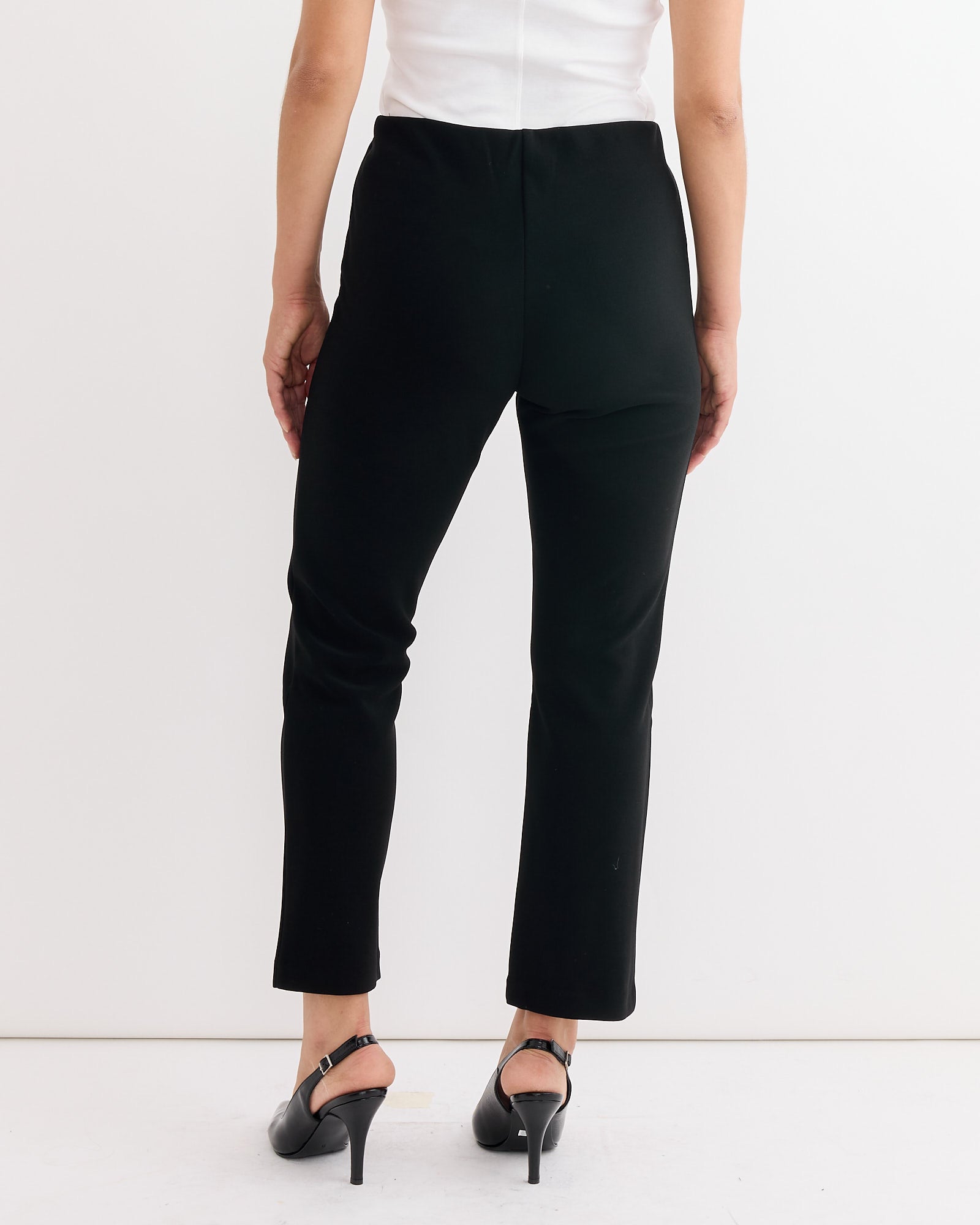 A woman faces away, wearing By Malene Birger's Vilea Cropped Trouser in Black, paired with a white top and black heels, against a plain white background.