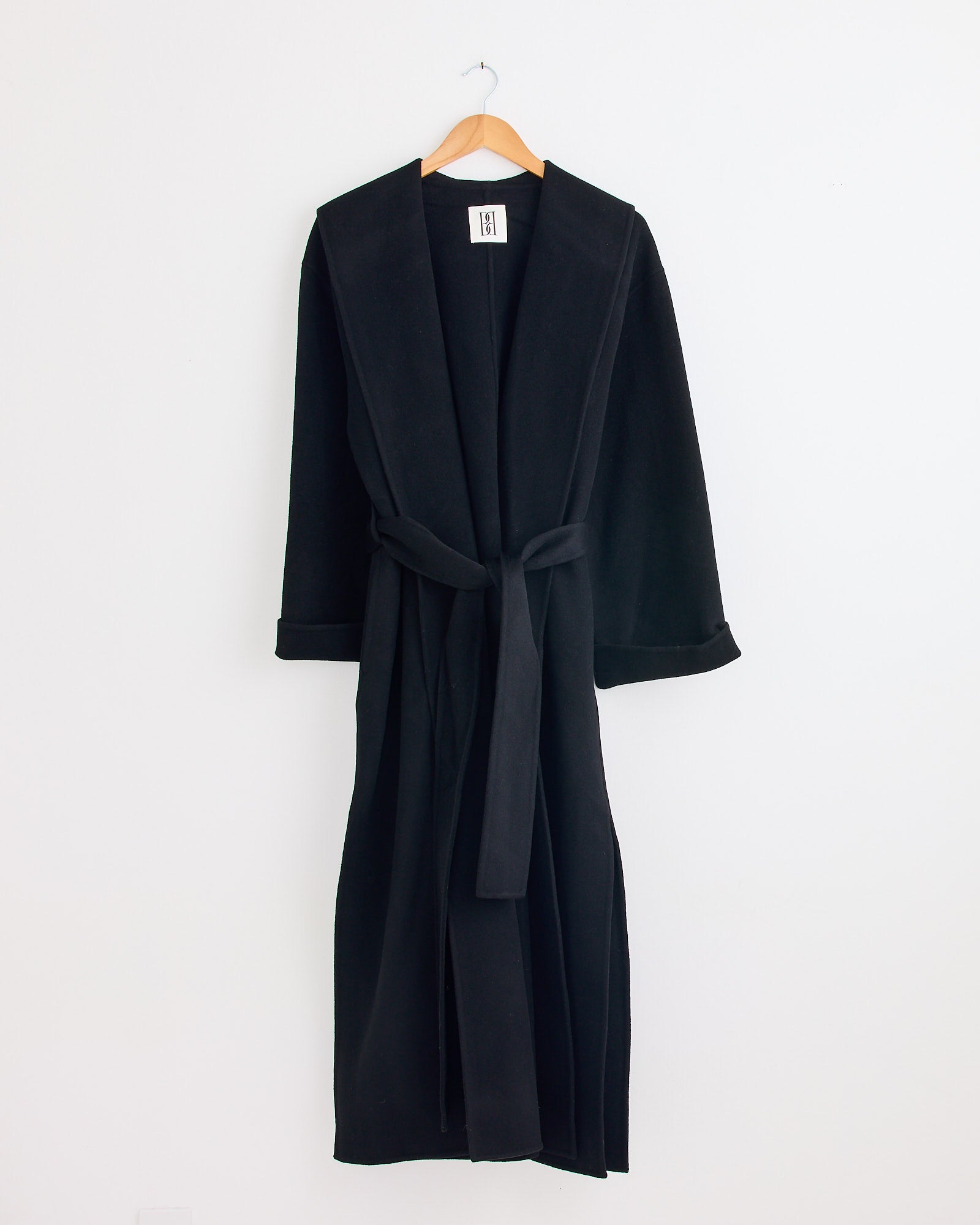 The Trullem Coat in Black by By Malene Birger hangs on a wooden hanger against a plain white wall—check sizing info or chat with us online to find your ideal fit.