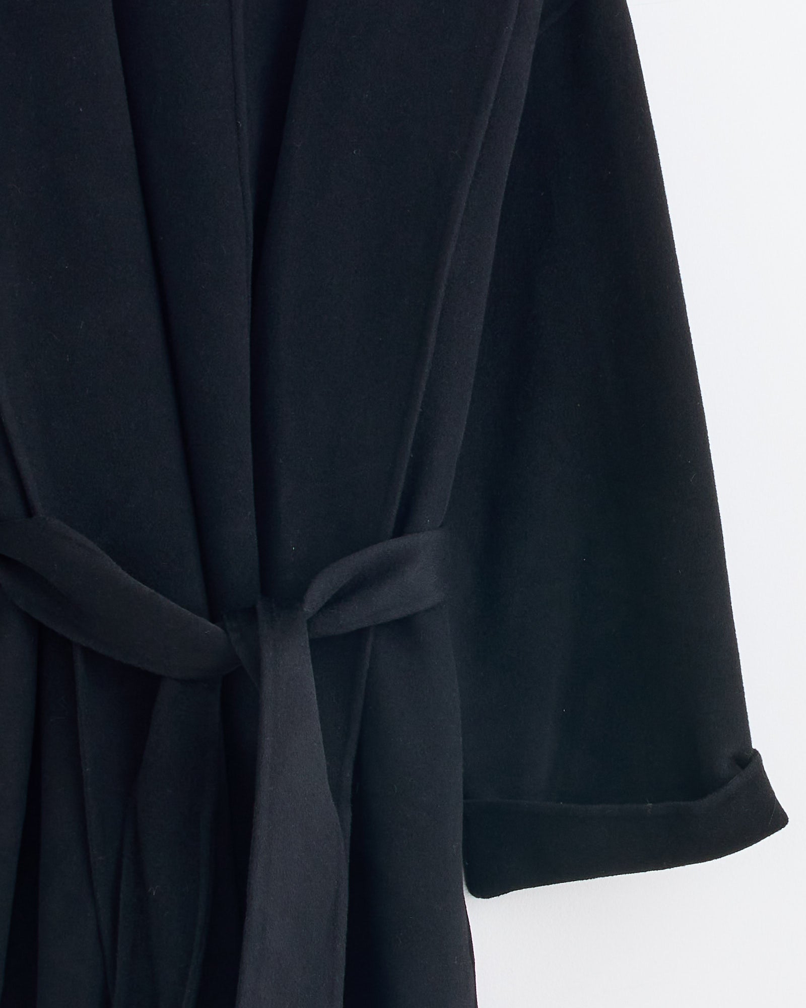 A close-up of the Trullem Coat in Black by By Malene Birger hangs against a white background, showcasing a fabric belt at the waist and sleeves with turned-up cuffs—perfect for achieving the ideal fit.