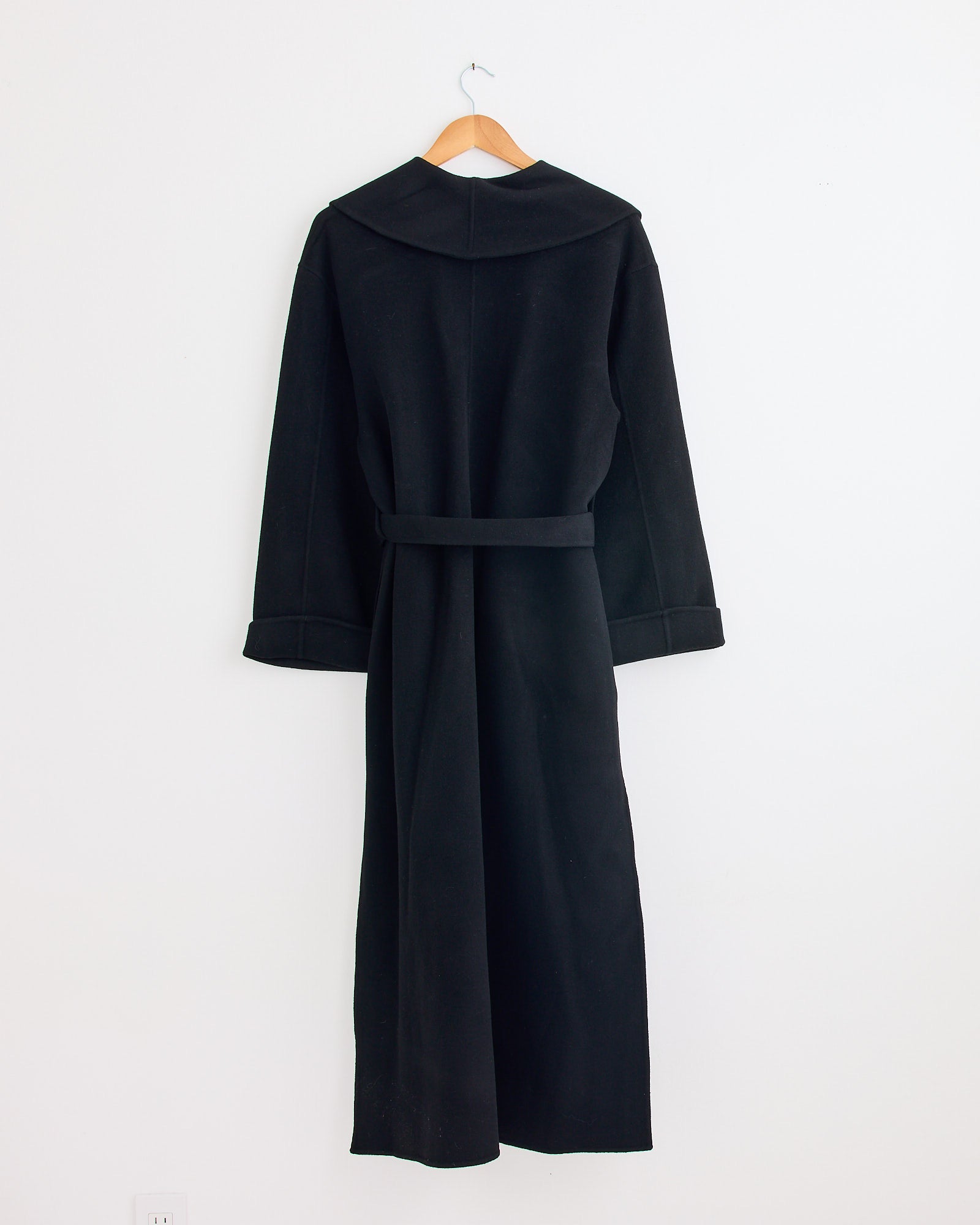 The Trullem Coat in Black by By Malene Birger, featuring a wide collar and tie belt, hangs on a wooden hanger against a plain white wall. For sizing or fit questions, contact us via online chat.