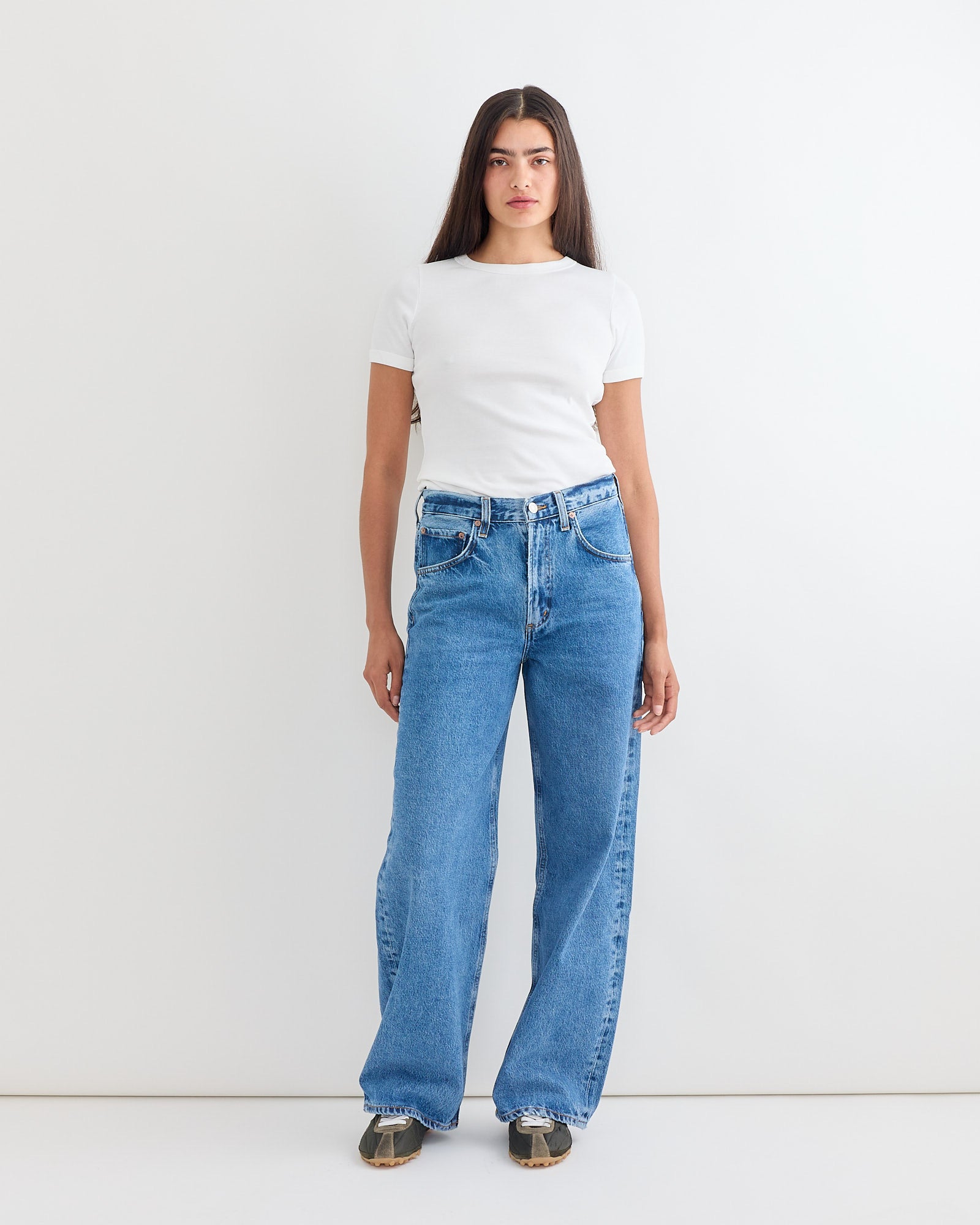 A woman with long straight brown hair wears the AGOLDE Low Curve Jean in Myth—high-waisted, loose blue jeans—paired with a fitted white short-sleeve tee and sneakers against a white background. Chat online for sizing or fit questions.