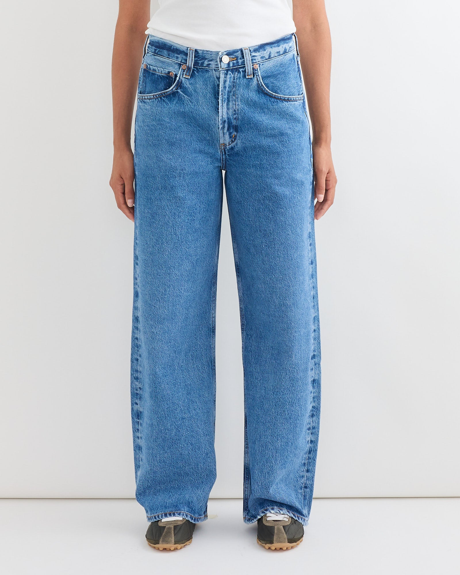 A person, shown from the shoulders down against a white background, wears AGOLDE’s Low Curve Jean in Myth styled with a tucked-in white shirt and dark sneakers, highlighting the jeans’ fit and sizing.