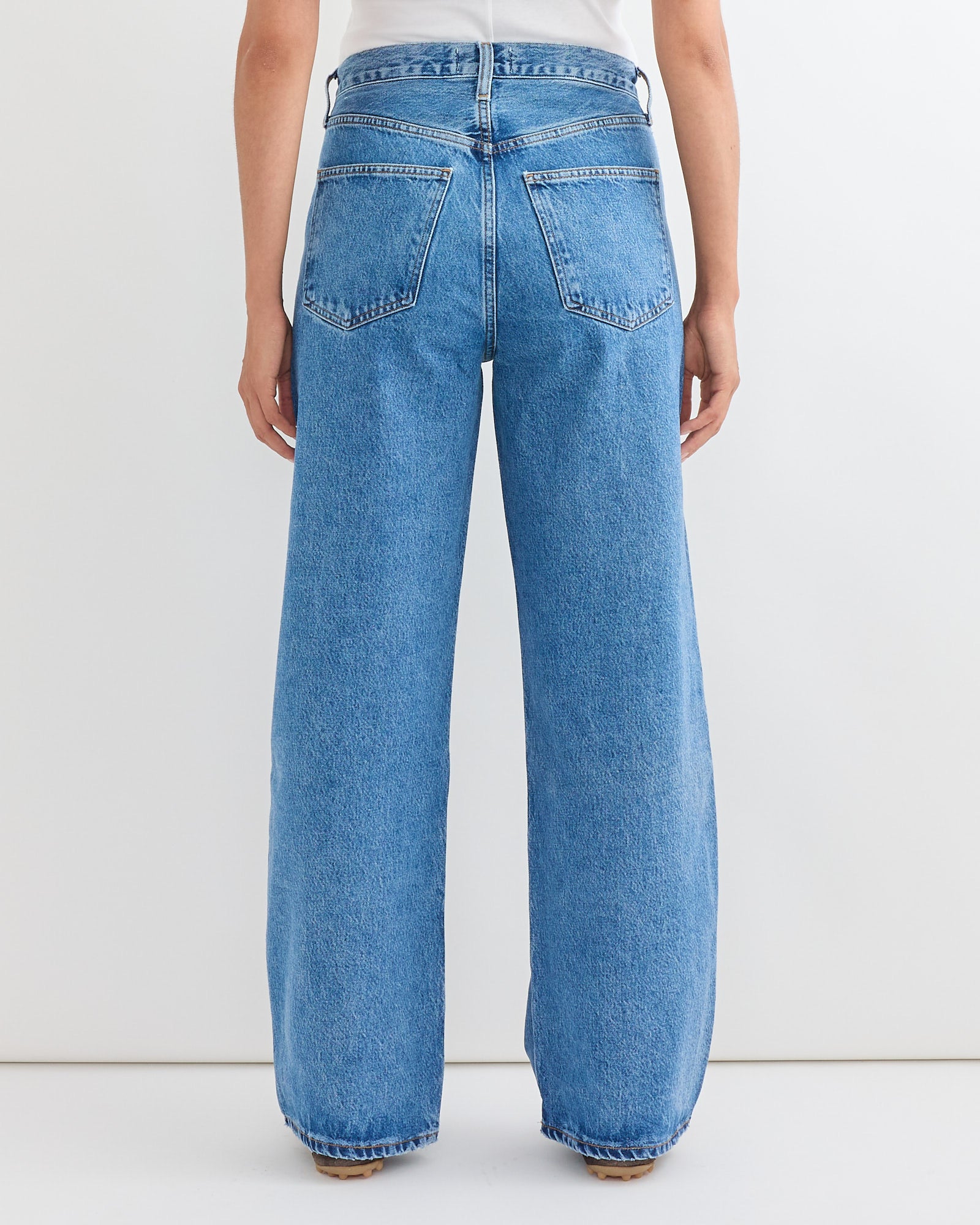 A person wearing AGOLDEs Low Curve Jean in Myth, a high-waisted light blue wide-leg style with a flattering fit, and a white top, shown from the back against a plain white background. Chat with us online for sizing help.